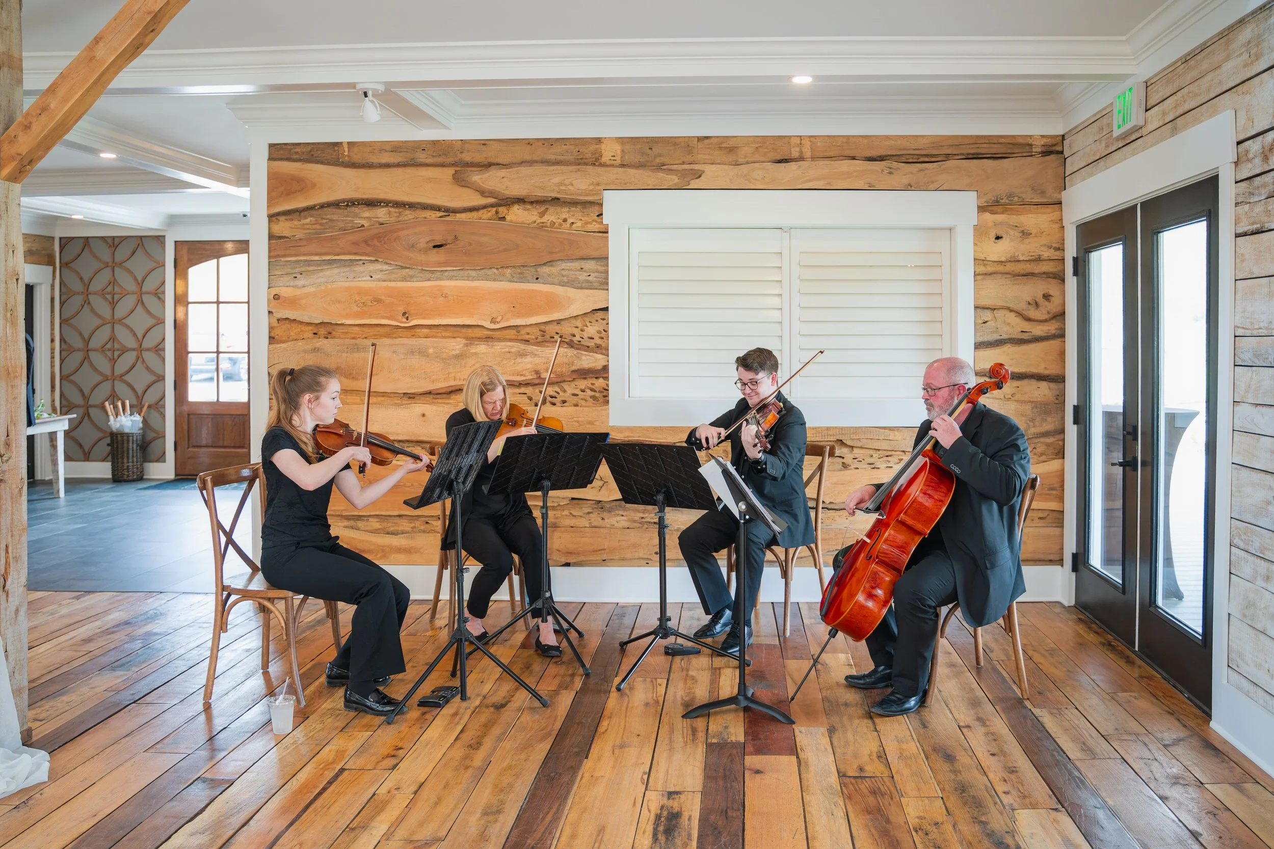 String quartet performing wedding ceremony music inside an elegant natural-vibe venue with wood floors, natural wood walls, black music stands, and large glass doors.