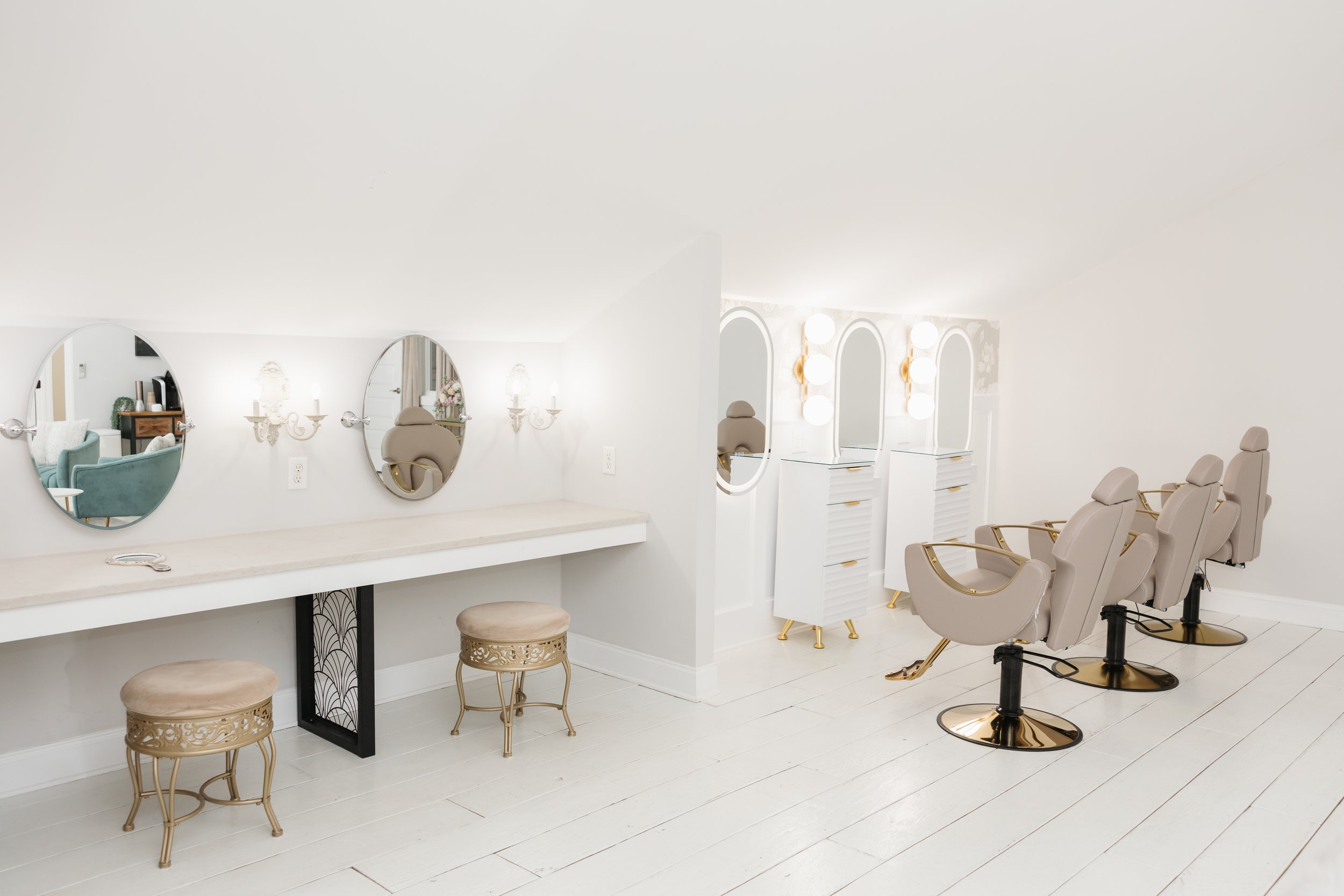 Bridal suite interior with white walls and flooring, featuring three mirrors, salon chairs with gold accents, vanity stations with lights, and small stools at Ramble Creek all-inclusive East Tennessee wedding venue.