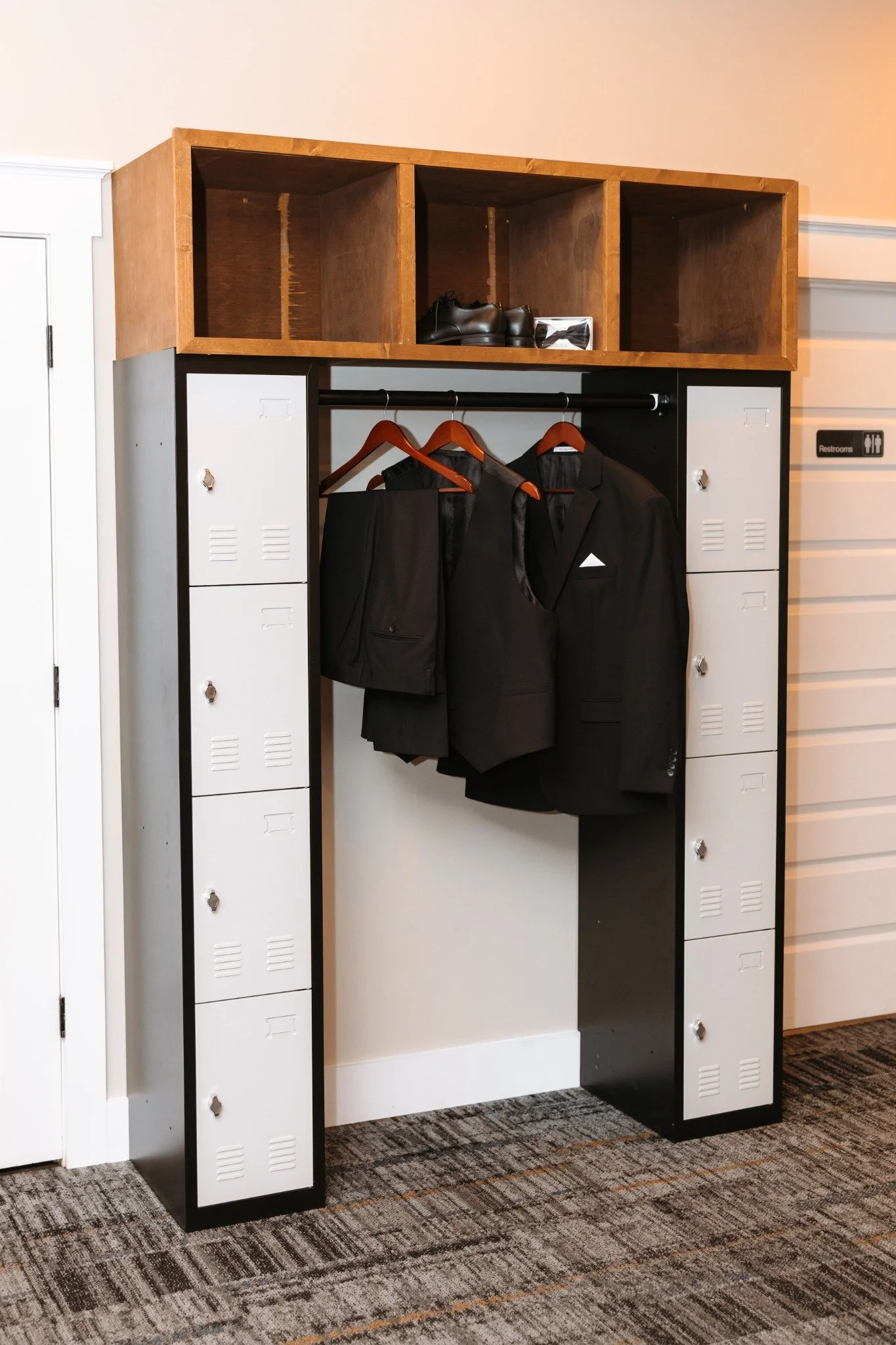 Locker unit with open cabinetry on top, hanging space with suits and jackets, shoes and accessories inside, located in an indoor space with carpeted floor and a restroom sign nearby, making the perfect place for grooms and their friends to get ready.