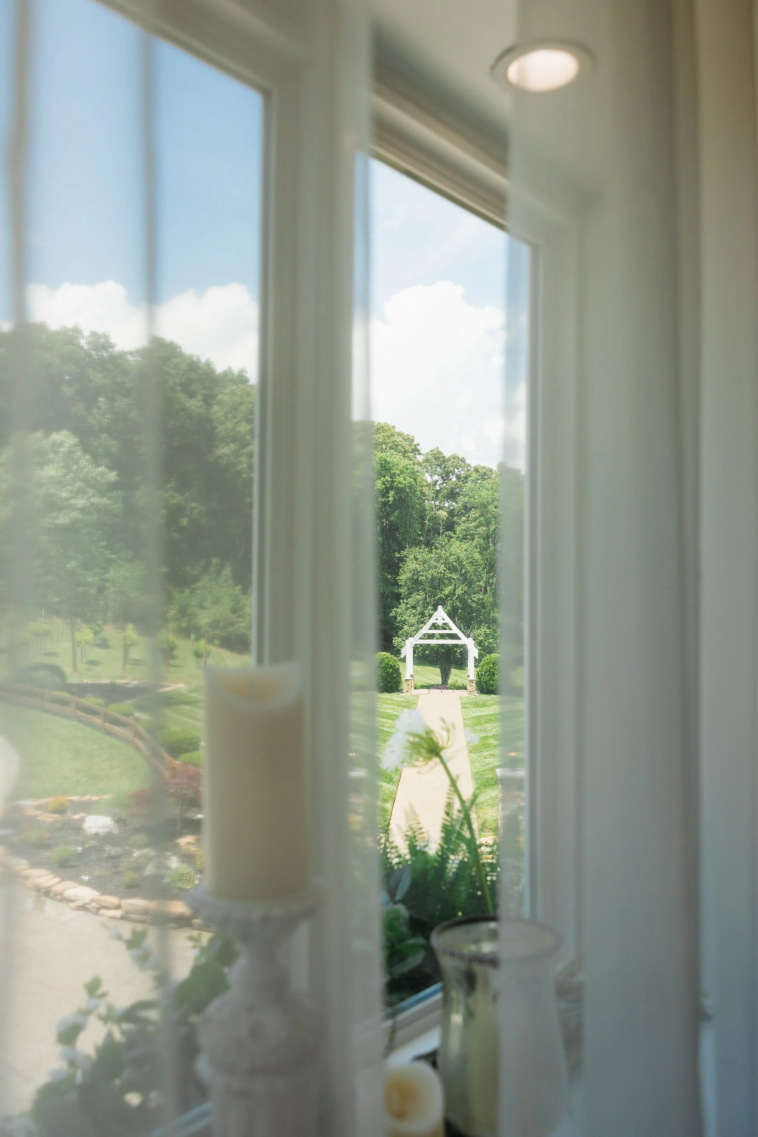 View through bridal suite window showing a green landscape where outdoor wedding ceremonies take place with trees and a blue sky with white clouds. Some objects, like candles and plants, on the window sill.
