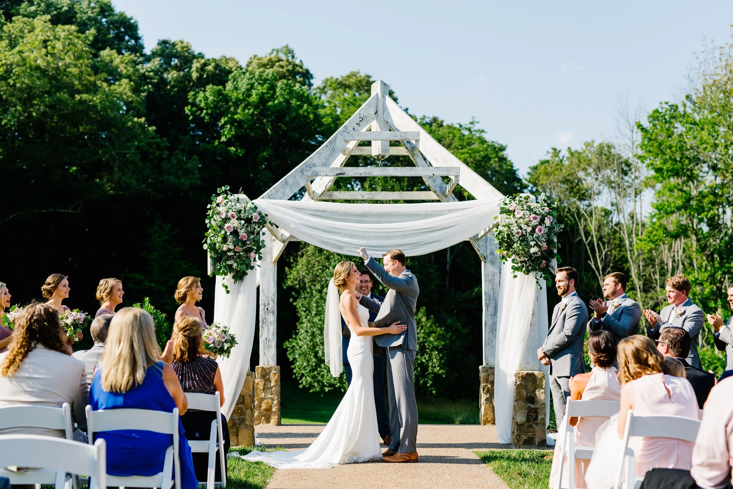 A couple exchanges vows during an outdoor wedding ceremony under a wooden arch decorated with flowers and draped fabric, with guests seated on white chairs and lush green trees in the background at Ramble Creek wedding venue in Athens, TN.