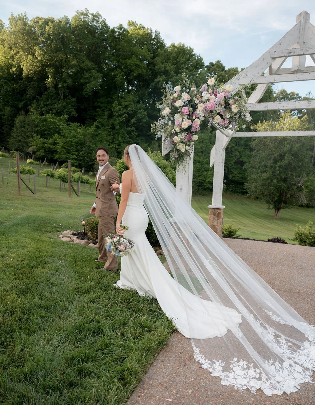 Bride and groom walking beneath the white outdoor wedding arbor at Ramble Creek all-inclusive wedding venue, with pastel spring florals, vineyard views, and the bride’s long lace veil trailing behind her