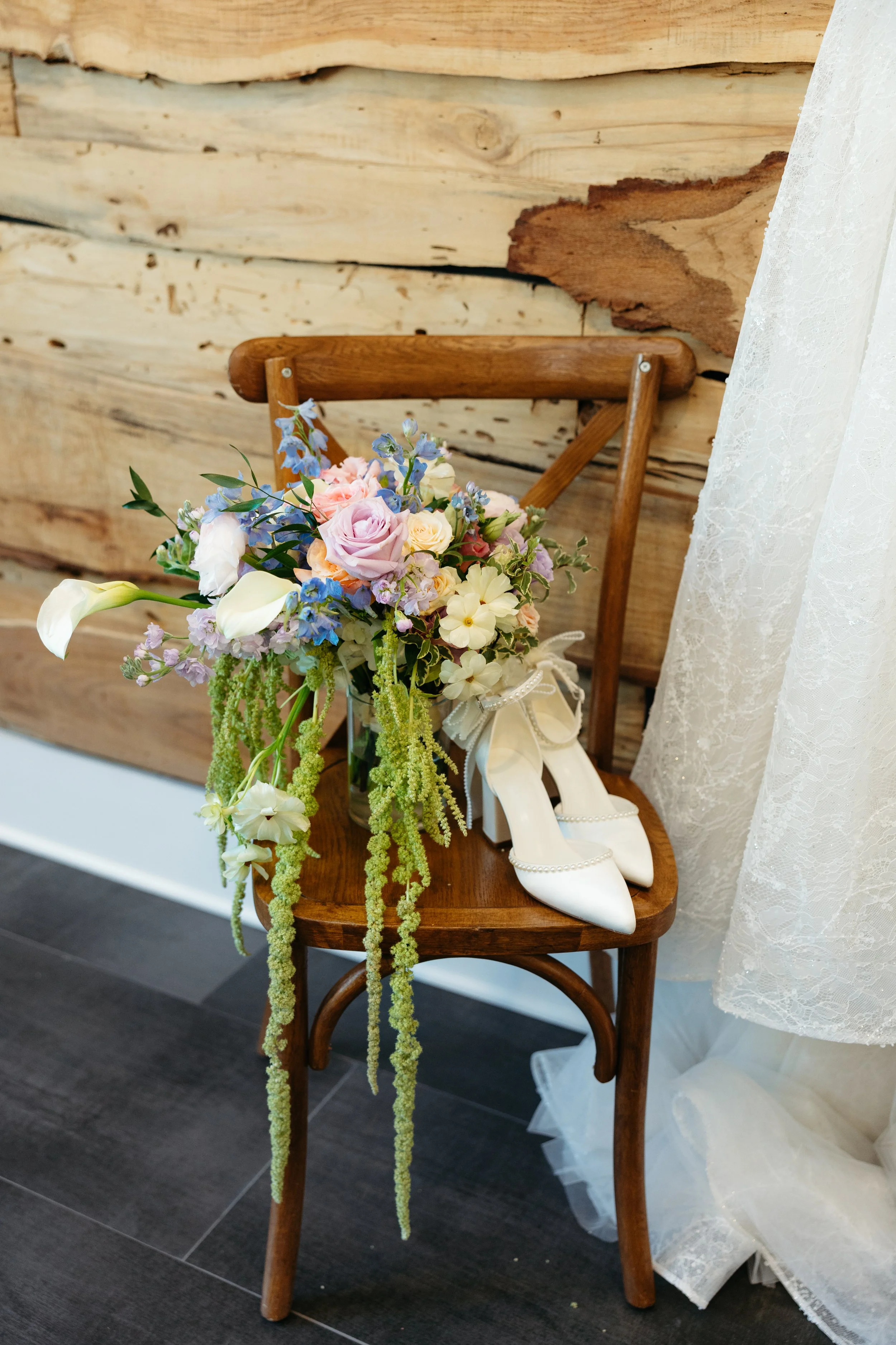 A wooden chair with a bridal bouquet of pastel-colored flowers and a pair of white high-heeled shoes on it, set against a rustic wood-paneled wall and wedding dress.