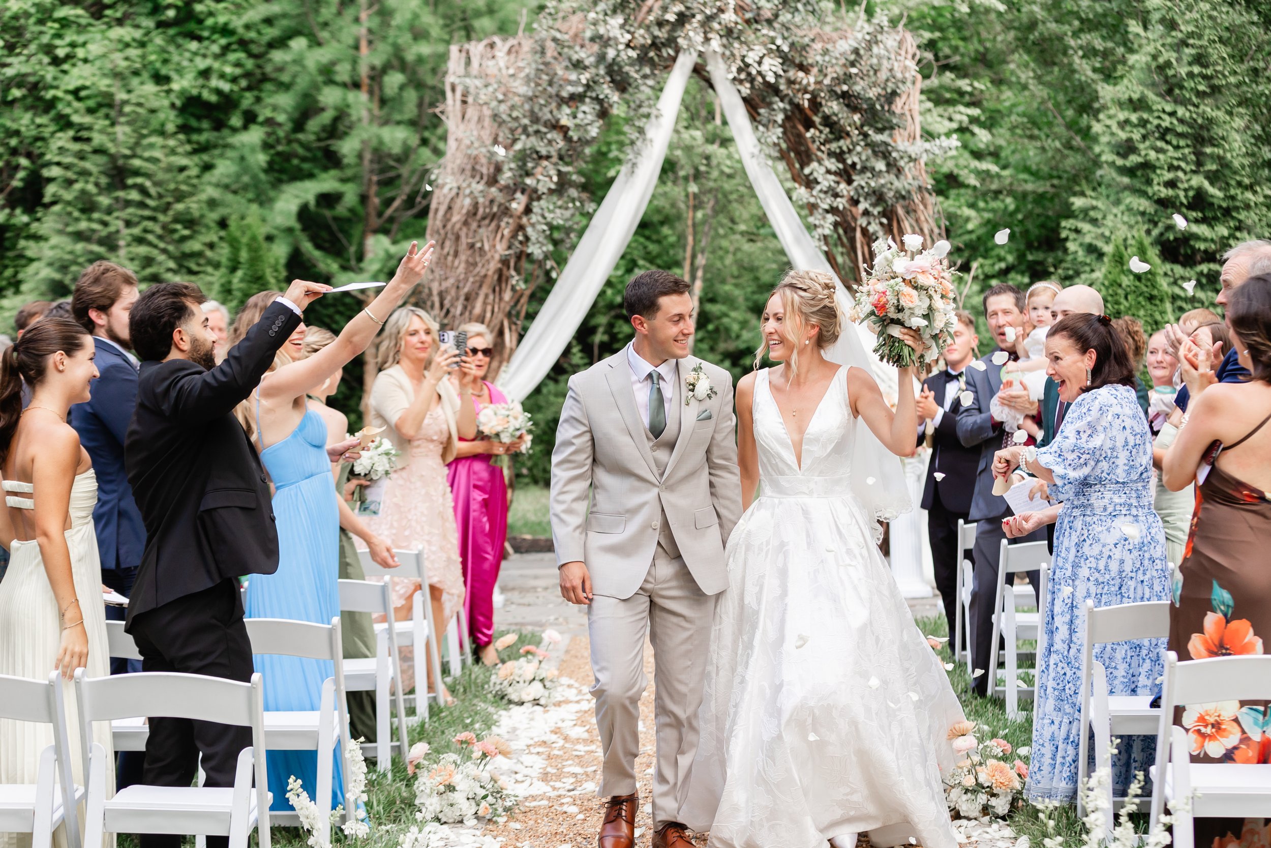 Bride and groom walk down the outdoor ceremony aisle at Ramble Creek as guests toss flower petals beneath the vine arbor.