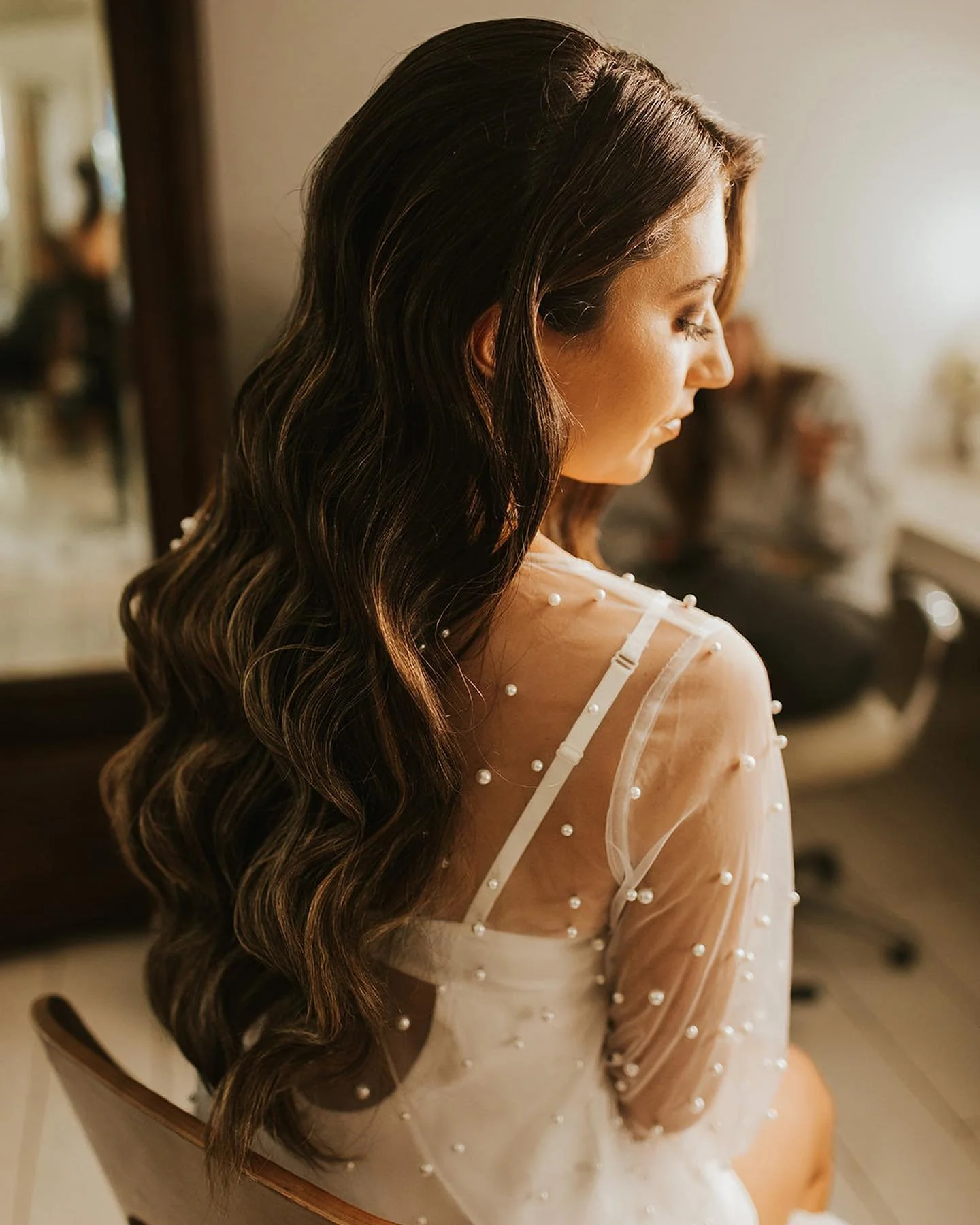 Bride with long, dark, wavy wedding hair seated in soft golden light, wearing a sheer pearl-dotted bridal top and looking down over her shoulder.