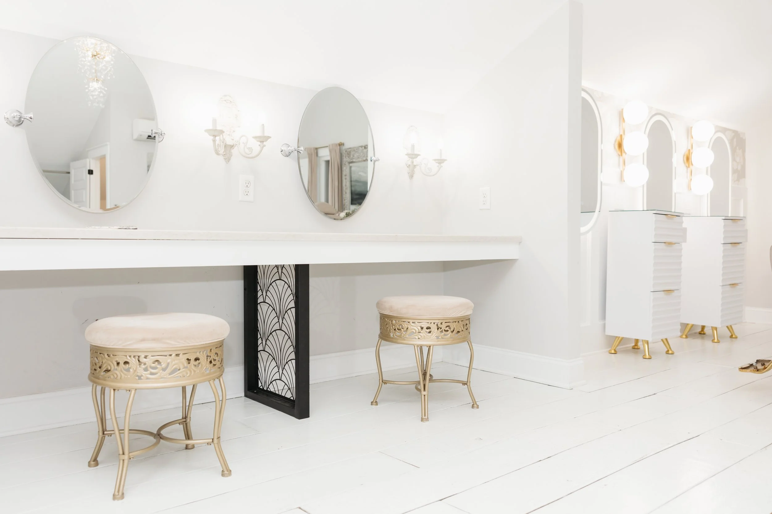 Bright, white modern interior with two oval mirrors, ornate wall sconces, beige stools, and a vanity with three glowing round lights where bridesmaids get ready on a bride's all inclusive wedding day at Ramble Creek.