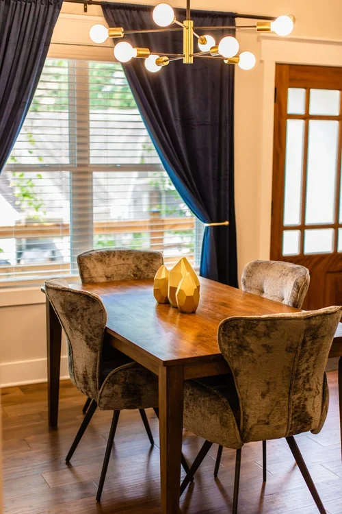 A dining room with a wooden table, four upholstered chairs, a modern chandelier, a window with blue curtains, and a wooden door. One of three accommodations at Ramble Creek Tennessee wedding venue with on-site lodging.