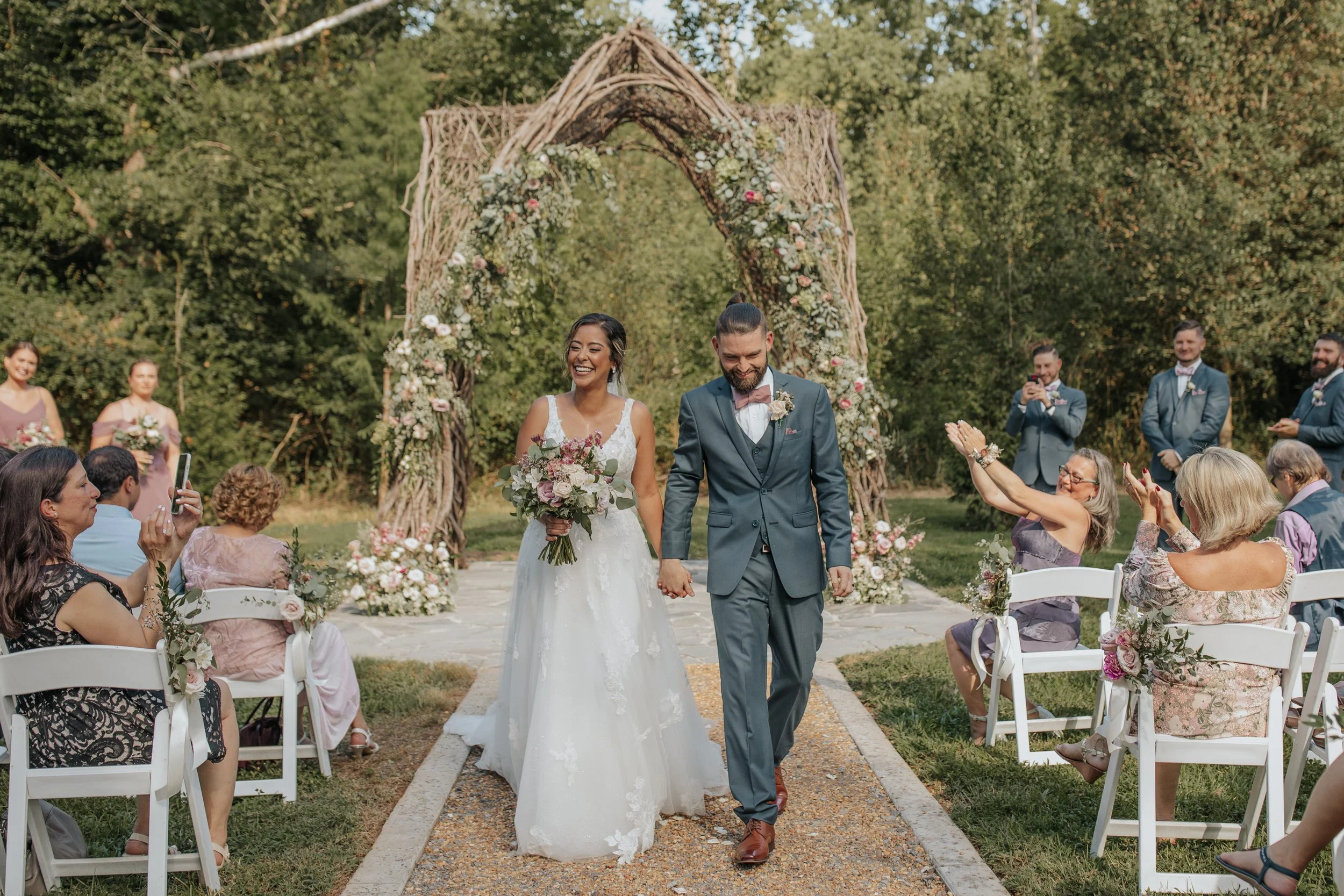 A bride and groom walk down the aisle outdoors at their wedding, surrounded by guests seated in white chairs decorated with flowers, with a floral arch behind them, in a green park setting.