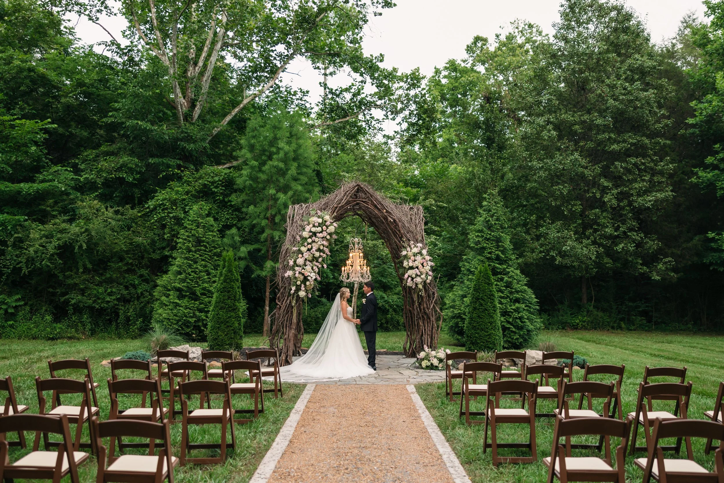 Outdoor woodland wedding ceremony at Ramble Creek Wedding Venue in Tennessee with bride and groom beneath a floral branch arch, chandelier, aisle, and guest seating.