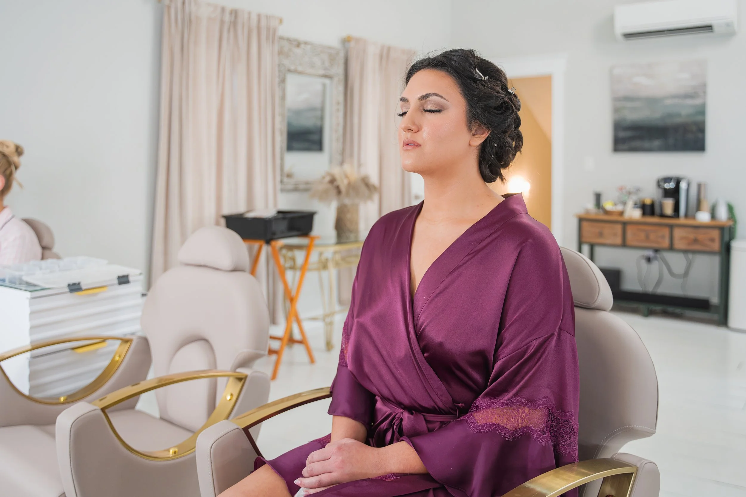 Bride sitting in a salon chair with eyes closed, showing soft bridal makeup and an elegant updo while wearing a plum satin robe
