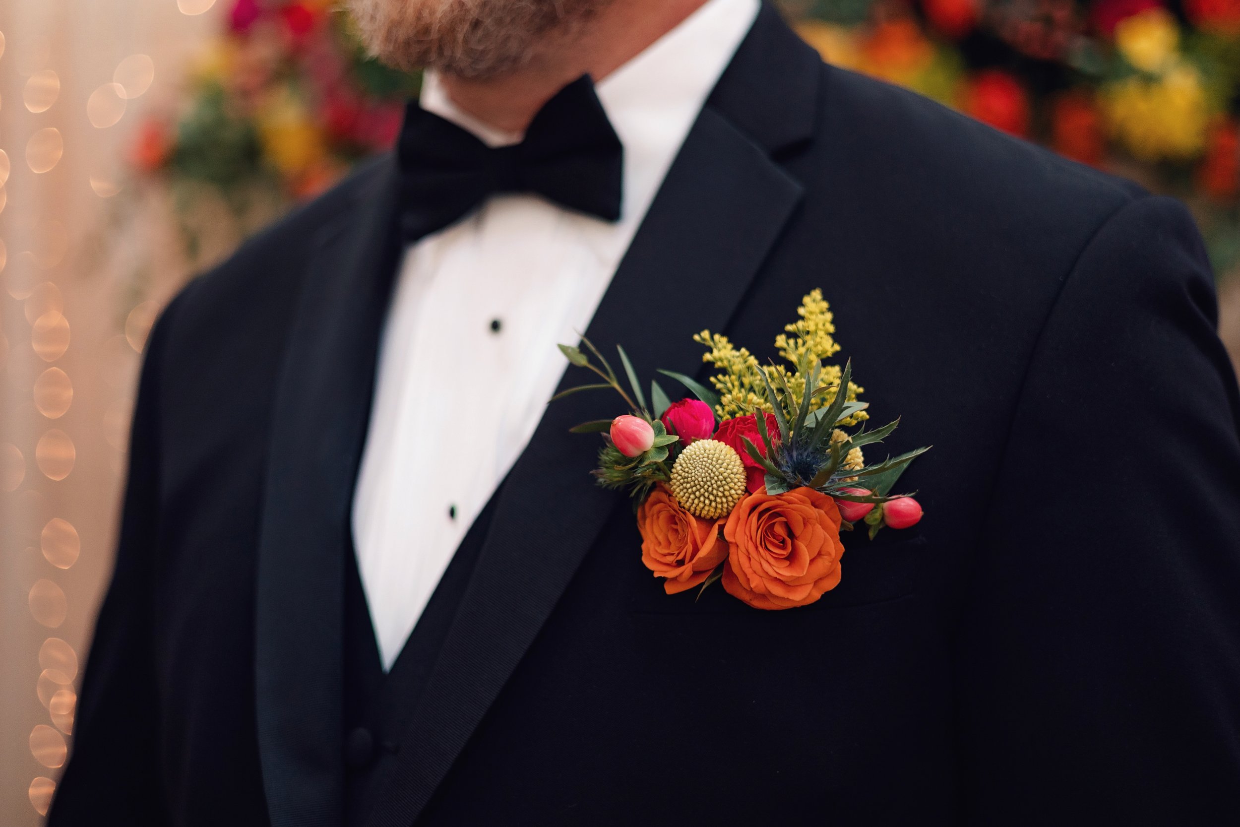 Close-up of a groom wearing a black tuxedo with a vibrant boutonniere featuring orange roses, yellow goldenrod, greenery, and berry accents at Ramble Creek.