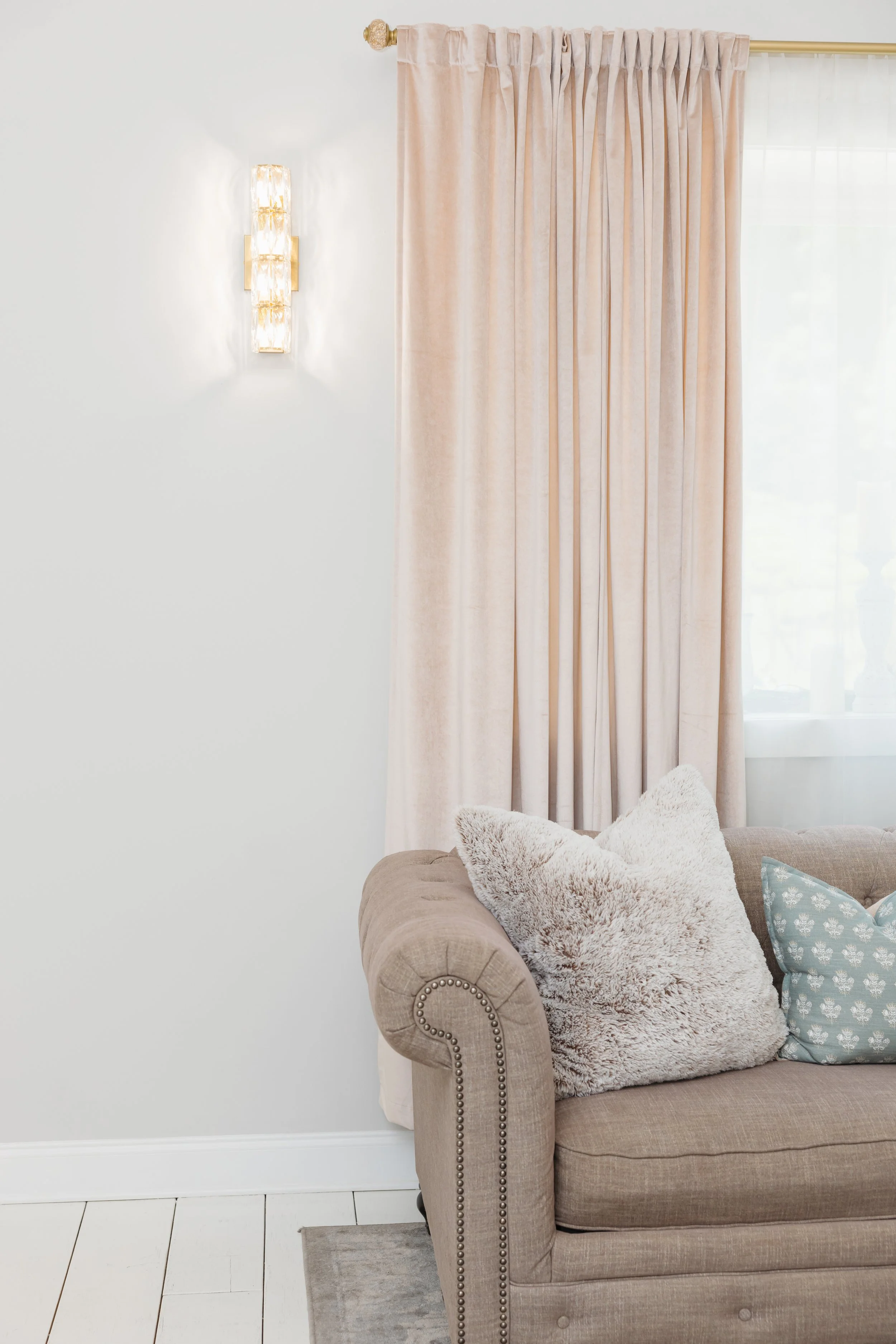 A beige upholstered sofa with decorative pillows, including a fluffy beige pillow and a blue patterned pillow, positioned against a white wall with cream curtains and a wall-mounted light fixture.