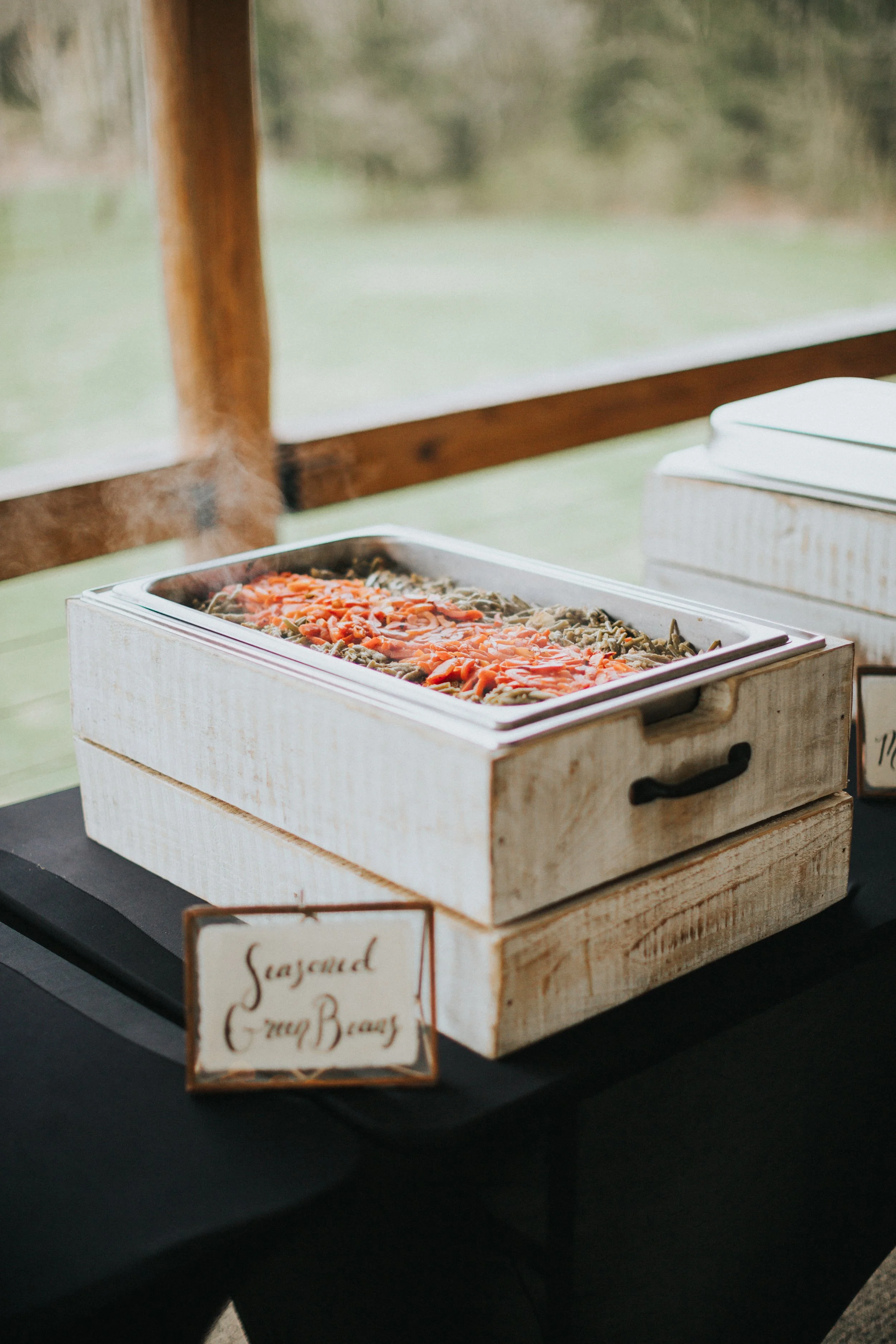 A wooden buffet service station with a metal lid holding cooked pasta topped with tomato sauce, situated on a black table at a wedding buffet, with a small labeled sign.