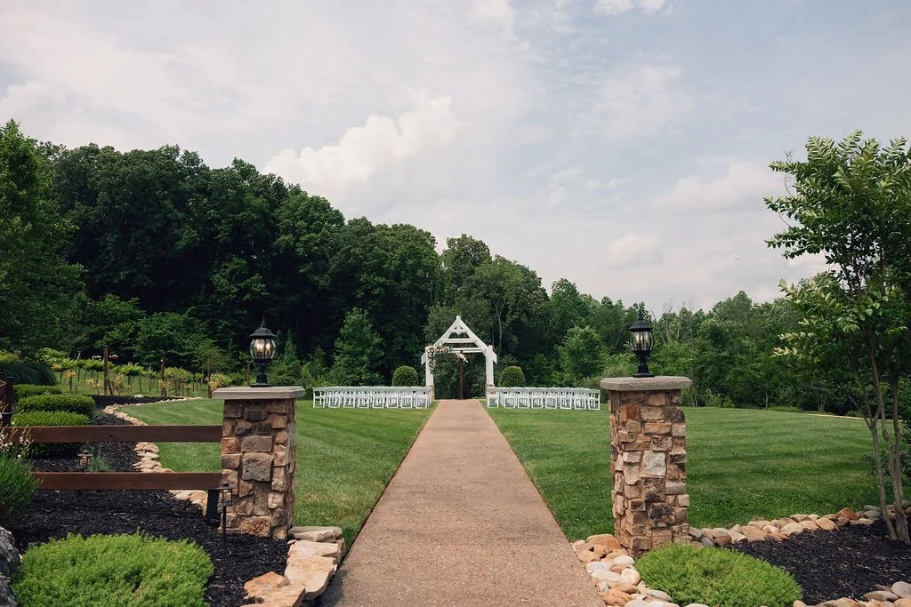A pathway leading to a wedding arch in a lush outdoor garden with neatly trimmed grass, trees, and rows of white chairs for guests at Ramble Creek all-inclusive wedding venue in Athens, TN.