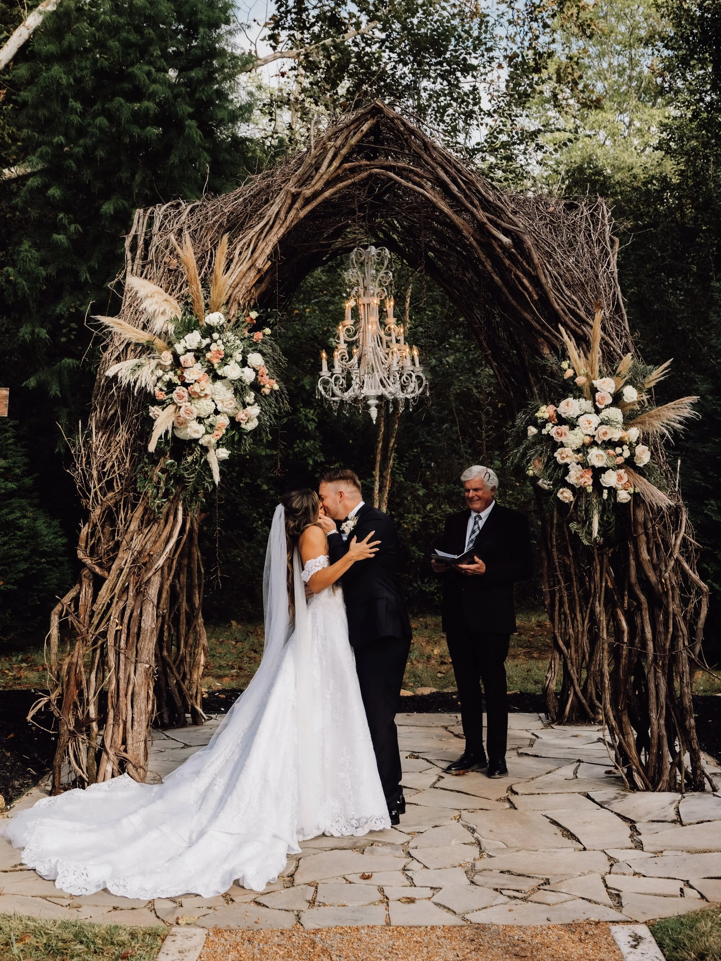 Kara Lewis and West Webb kiss beneath Ramble Creek’s Vine Arbor, framed by blush and ivory flowers, pampas grass, and a chandelier during their outdoor wedding ceremony