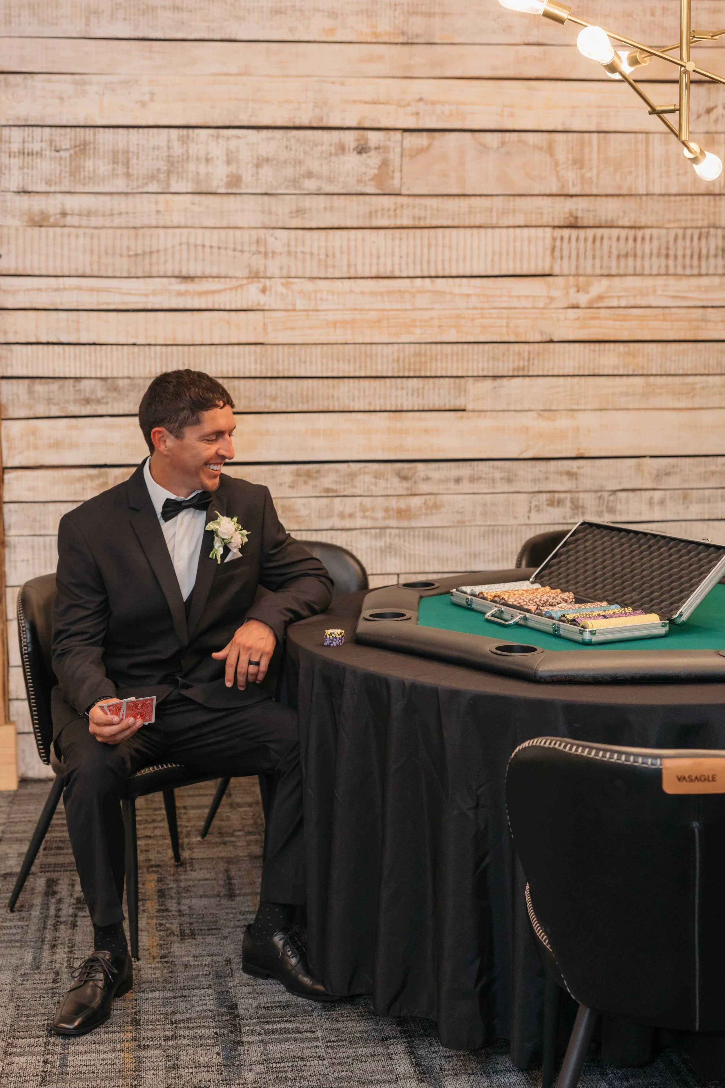 Groom in black tuxedo plays poker while getting ready for his wedding day at Ramble Creek wedding venue, with spacious and private groom's quarters.