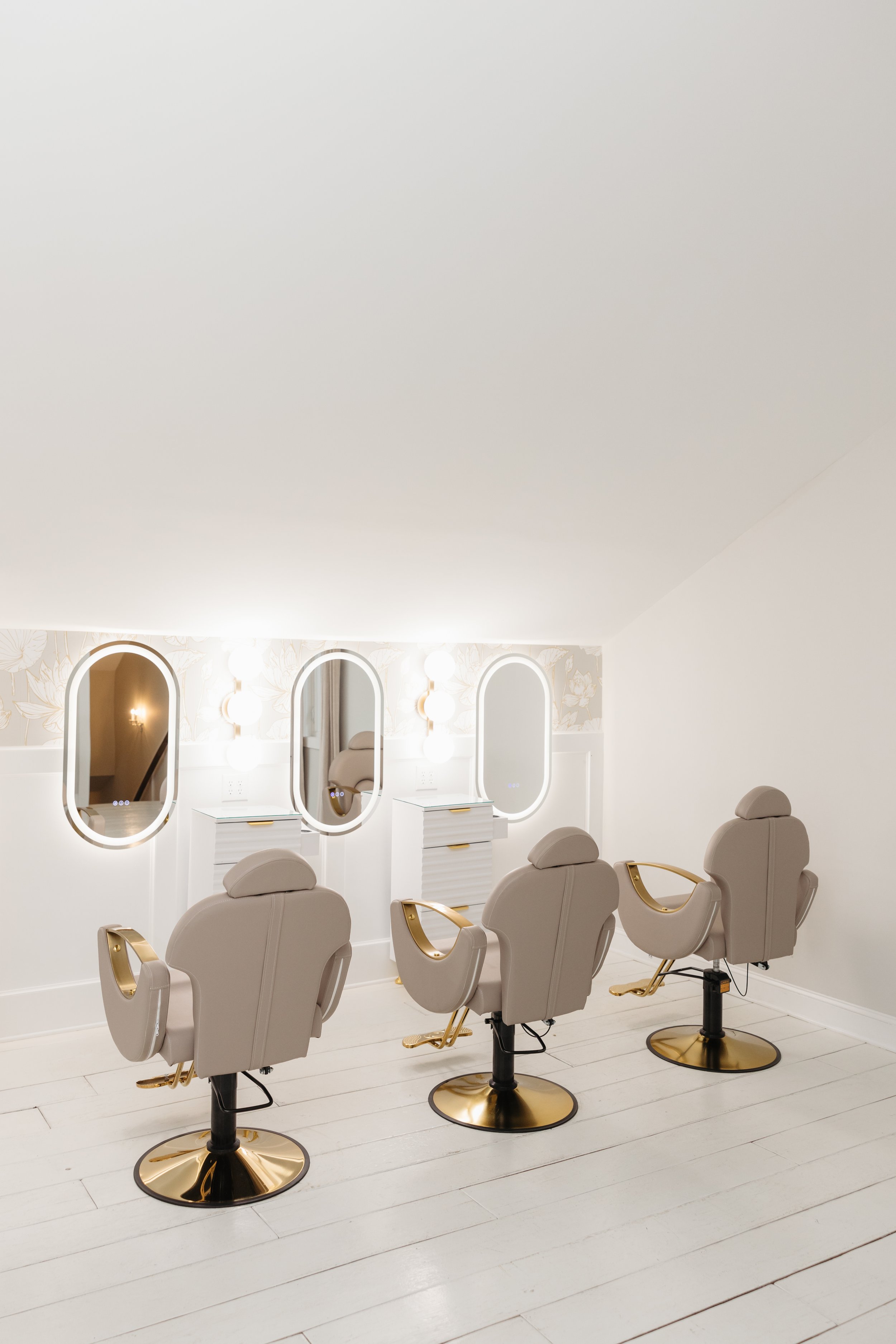 A modern getting ready space for Ramble Creek brides with four beige styling chairs with gold accents positioned in front of three large oval mirrors mounted on a white wall with a floral wallpaper border. The floor is white wooden planks.
