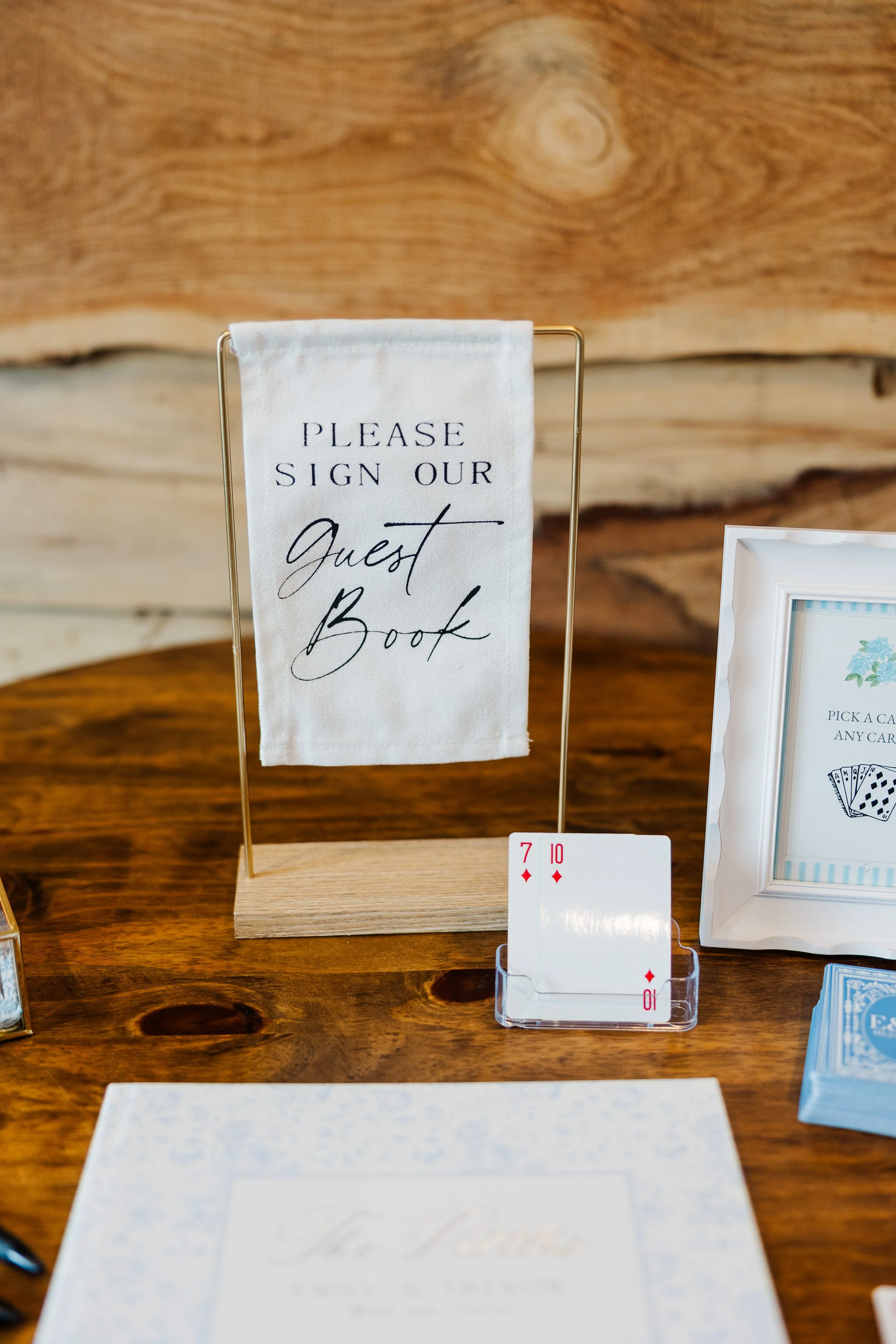 Wedding guest book display at Ramble Creek Wedding Venue in Tennessee featuring a white fabric sign, playing cards, and elegant reception details on a wood table.