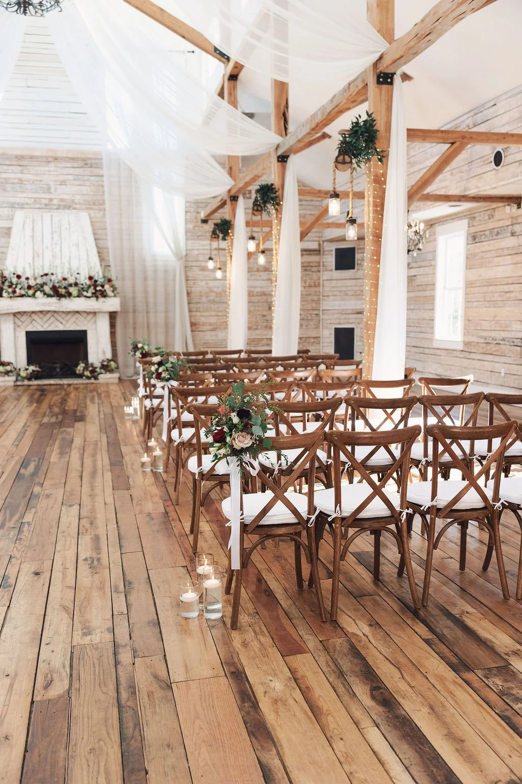 Interior of a beautiful wedding ceremony space with wooden chairs decorated with flowers, a stone fireplace with floral arrangements, and hanging lanterns, with white drapes and string lights hanging from wooden beams.