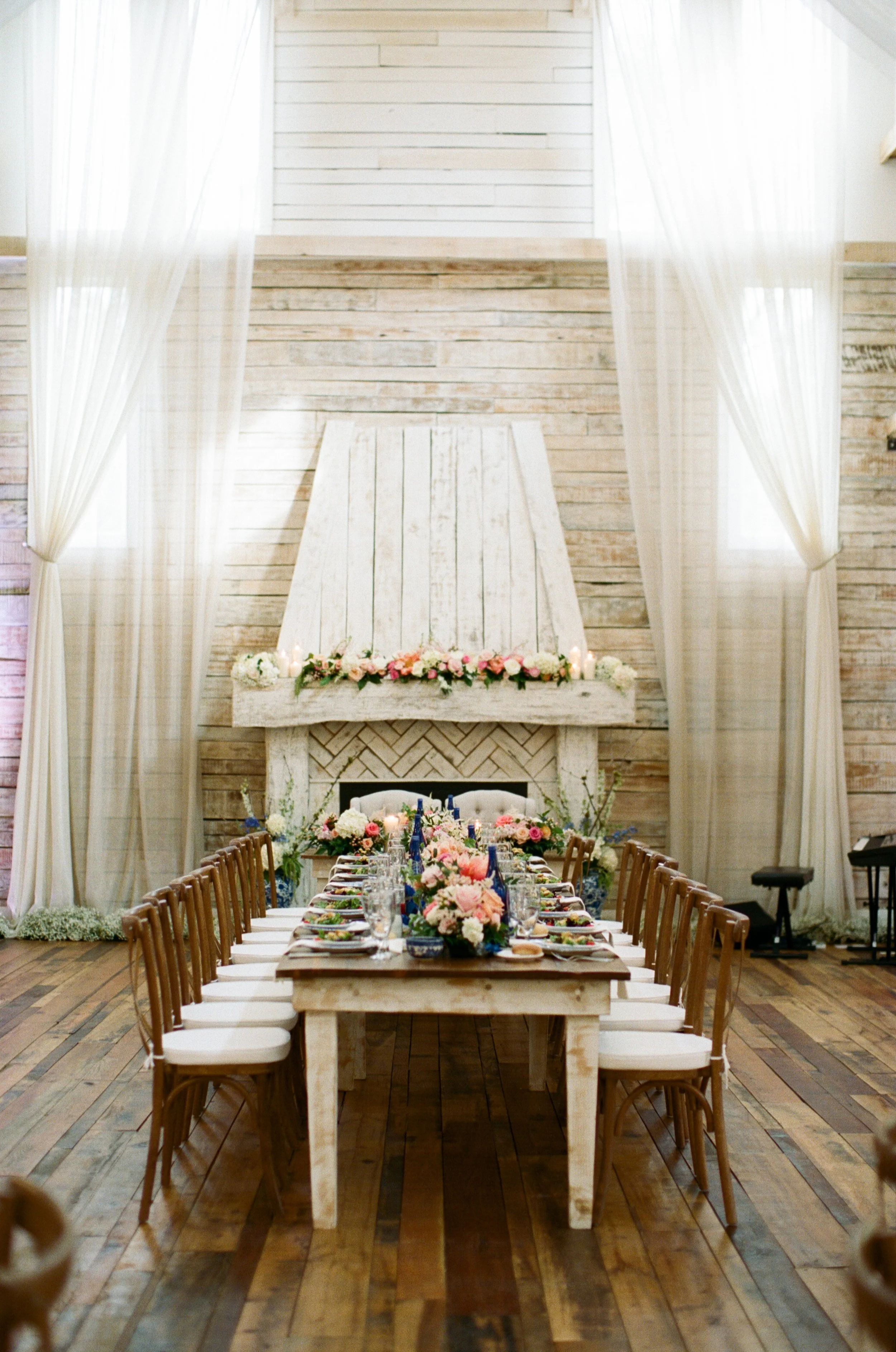 Long reception table with romantic floral centerpieces featuring blush roses, blue delphinium, greenery, and candlelight arranged inside the Grand Hall at Ramble Creek, framed by soft drapery and warm wood details.