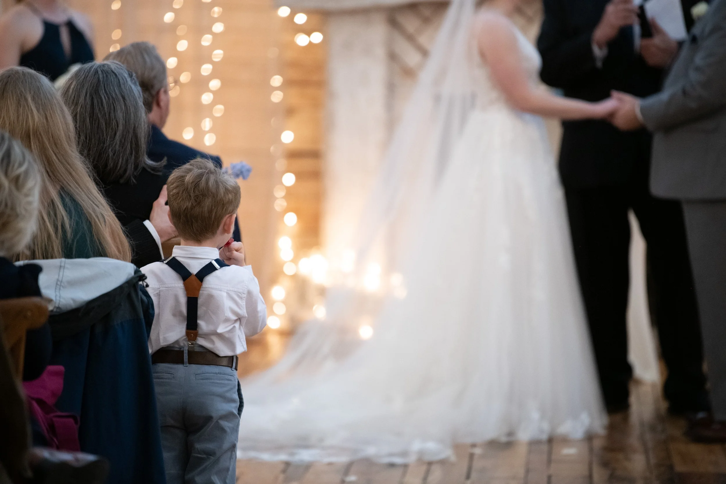 Guests seated at an indoor Knoxville-area wedding ceremony, focused on the couple exchanging vows, with wedding decorations and string lights in the background.