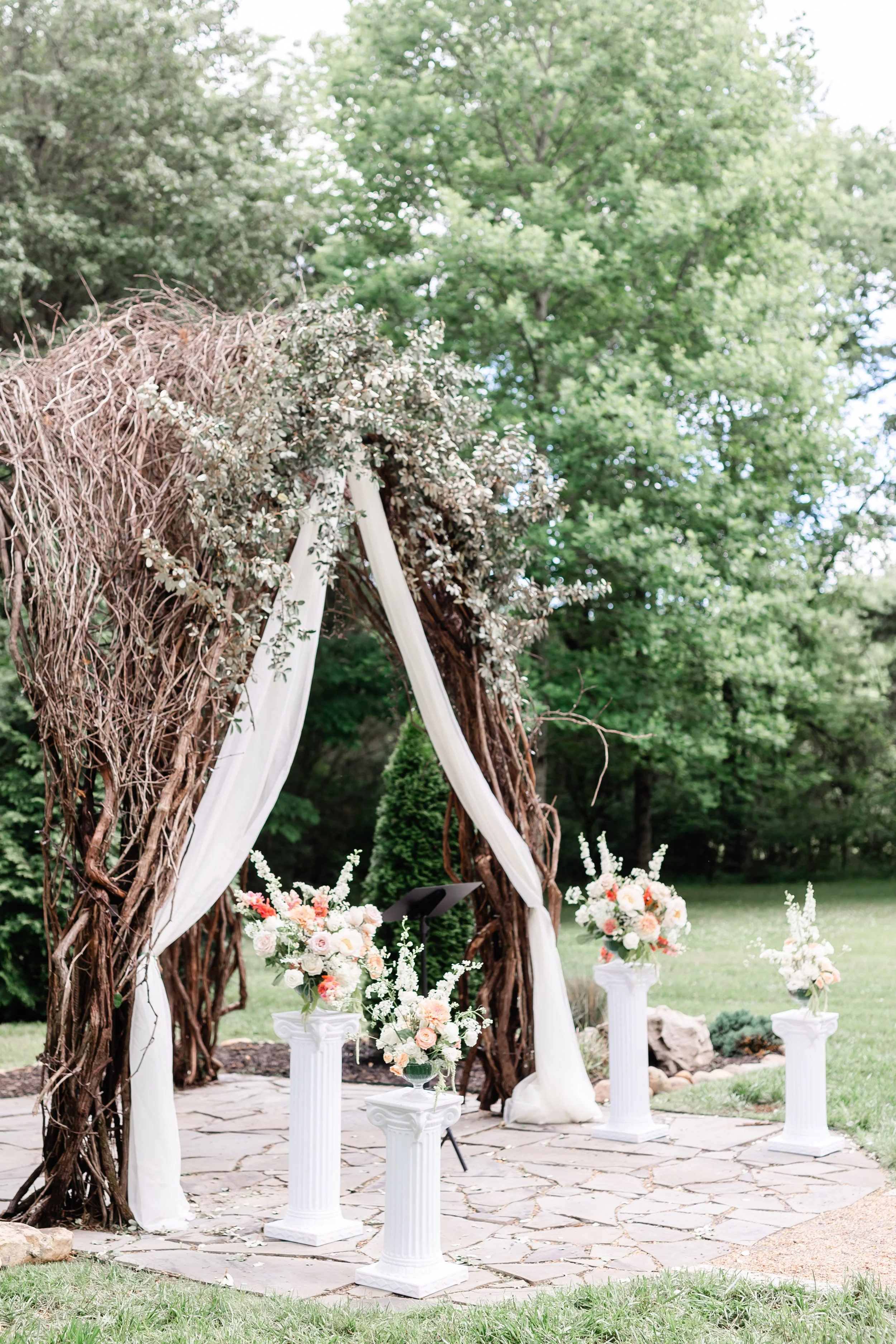 Outdoor wedding arch in Tennessee decorated with white fabric, Muscadine vines, and floral arrangements, set on a stone patio with floral arrangements in white pedestals, greenery, and trees in the background.