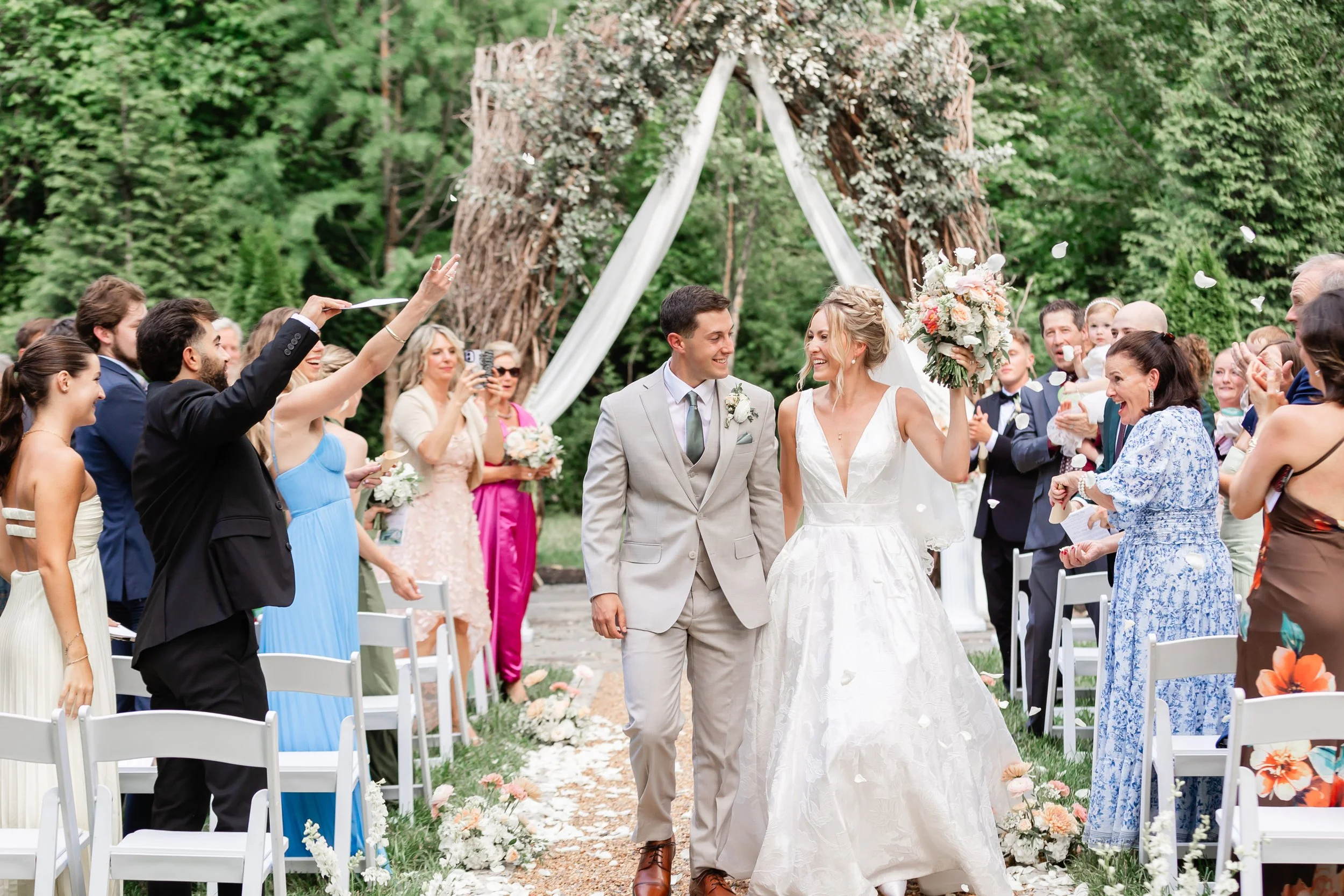 Newlyweds walk down the aisle after their outdoor wedding ceremony at Ramble Creek’s vine arbor, surrounded by cheering guests, white chairs, lush floral aisle decor, and wooded Tennessee scenery