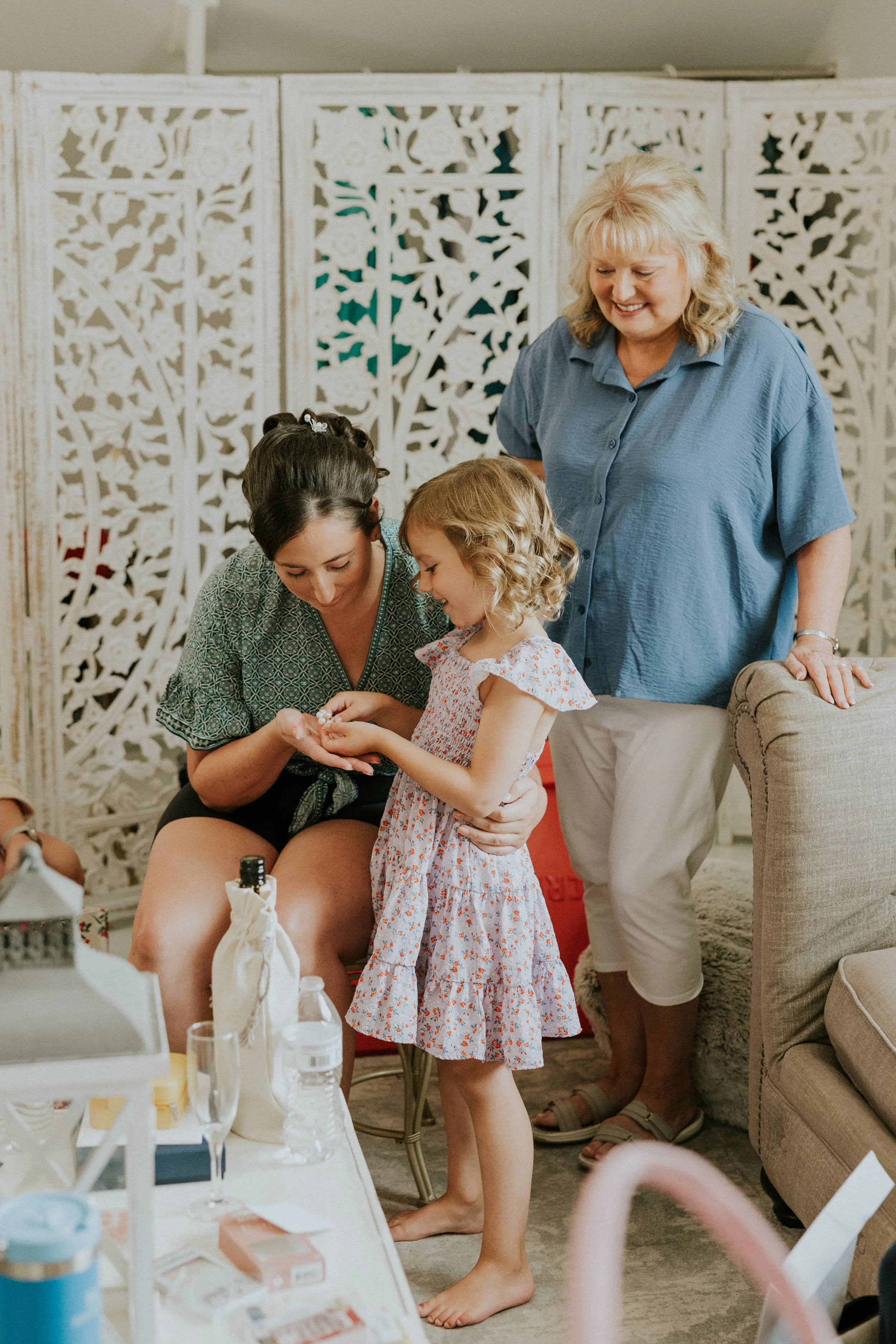 A young girl with curly blonde hair in a floral dress receiving an item from a woman seated, with multiple women and a woman in a blue shirt observing in the background, set in the Ramble Creek bridal suite near Chattanooga.