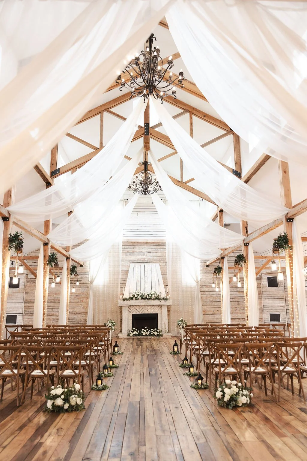 A rustic but elegant wedding ceremony setup with wooden chairs, white floral arrangements, hanging lanterns, and flowing white drapery inside a light-filled Grand Hall with wooden beams and chandeliers.