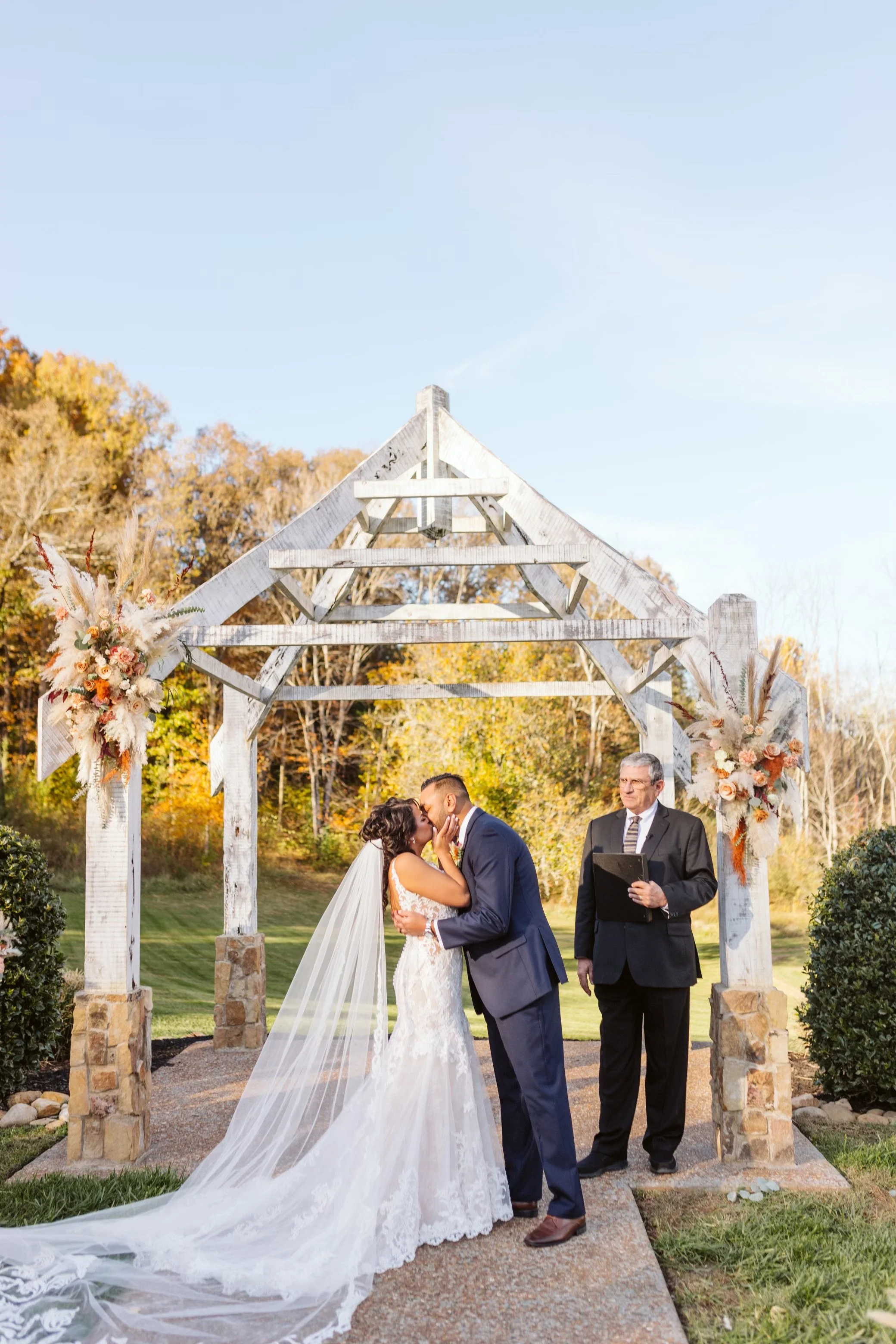 Outdoor wedding ceremony with a bride and groom kissing under a wooden arch decorated with flowers. The bride is in white gown with a long veil, and the groom in a navy suit. An officiant stands to the side holding a folder, with fall trees.