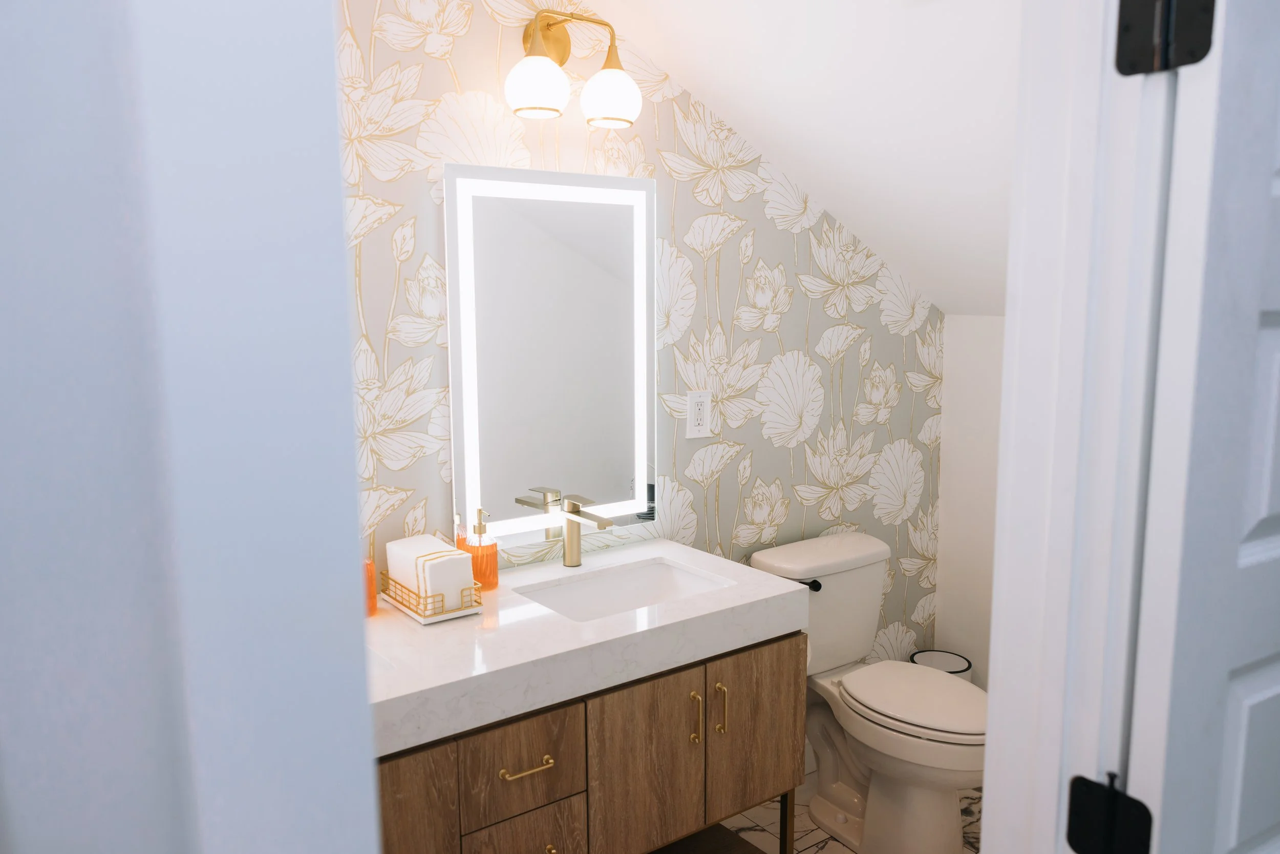Ramble Creek private bridal suite bathroom with floral wallpaper, illuminated mirror, gold light fixtures, countertop with soap dispensers and tissues, and a toilet.