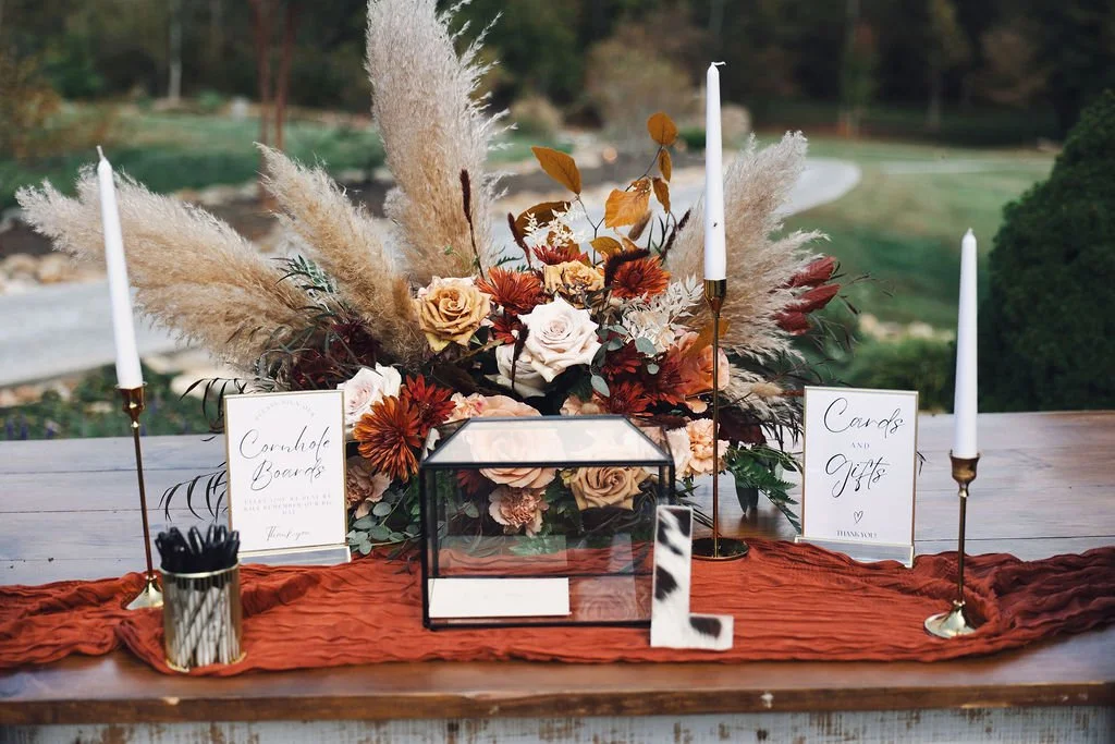 Fall wedding welcome table with rust-colored gauze runner, glass card box, taper candles, and boho floral arrangement featuring pampas grass, roses, autumn leaves, and warm orange blooms.