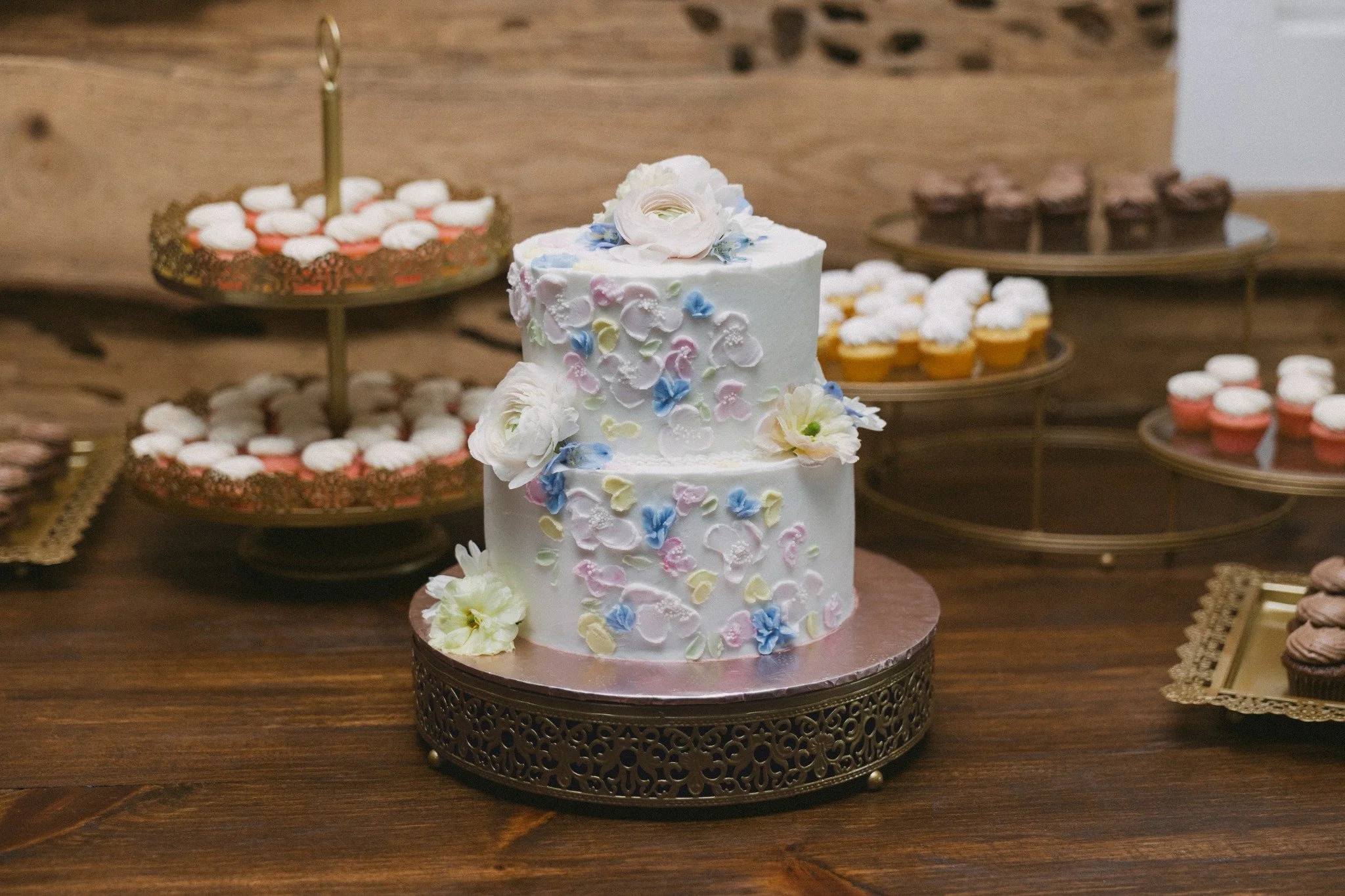 Floral two-tier wedding cake surrounded by mini desserts on a Ramble Creek reception dessert table.