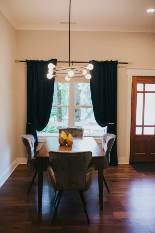Dining room with dark curtains, a wooden table with three upholstered chairs, a modern chandelier, a window with trees outside, and a wooden door. One of three accommodations at Ramble Creek Tennessee wedding venue with on-site lodging.