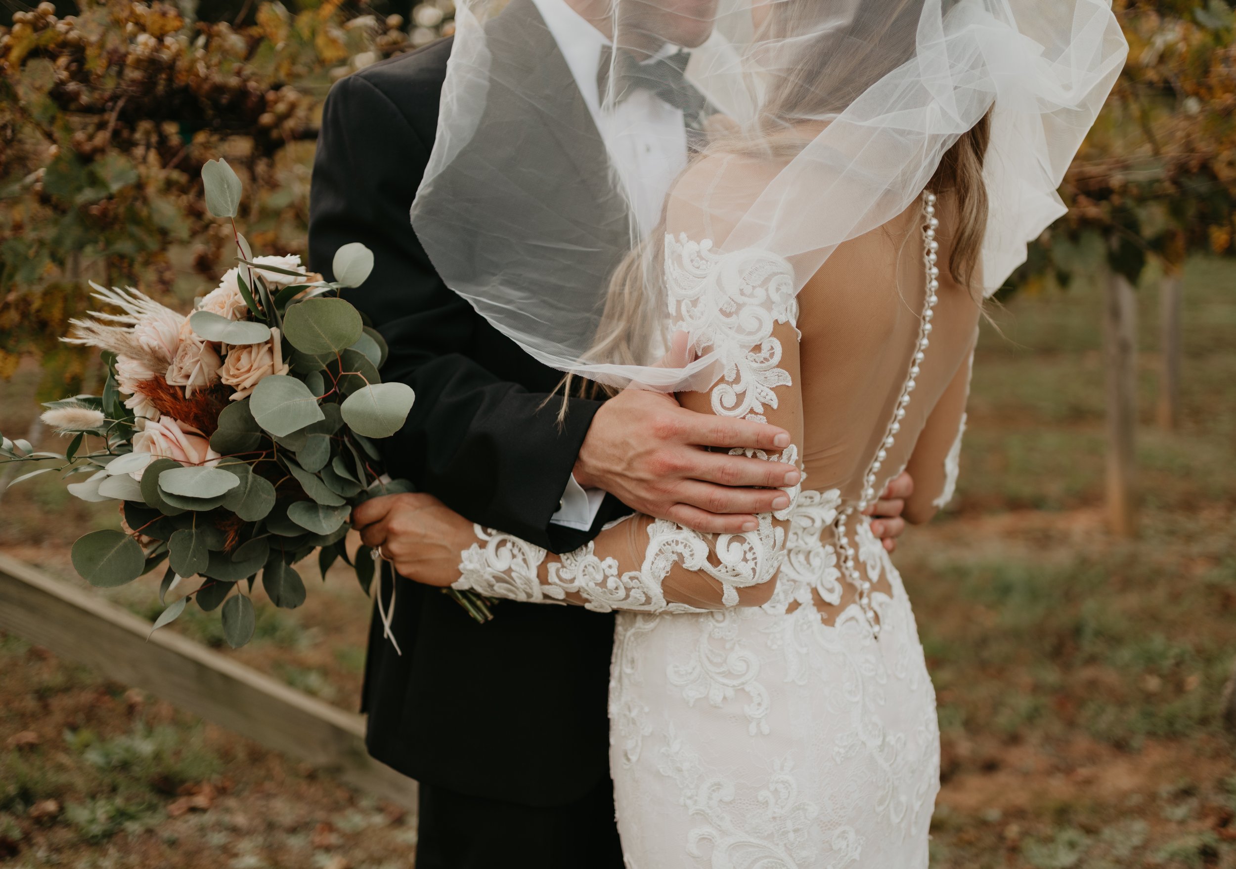 Bride and groom embracing in Ramble Creek vineyard, with the bride holding a soft neutral bouquet and wearing a lace wedding dress with pearl button details
