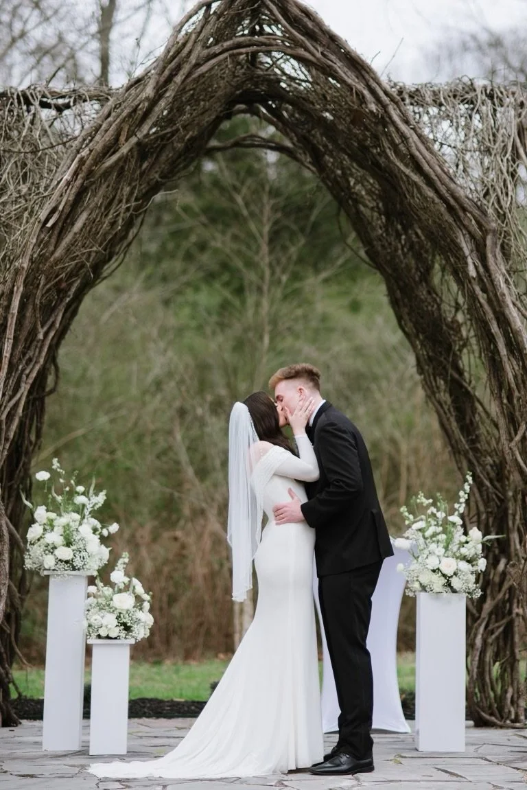 A bride and groom share a kiss during their outdoor wedding ceremony under a large arch made of intertwined branches. The bride wears a white wedding dress and veil, while the groom wears a black suit. There are white floral arrangements on tall pede