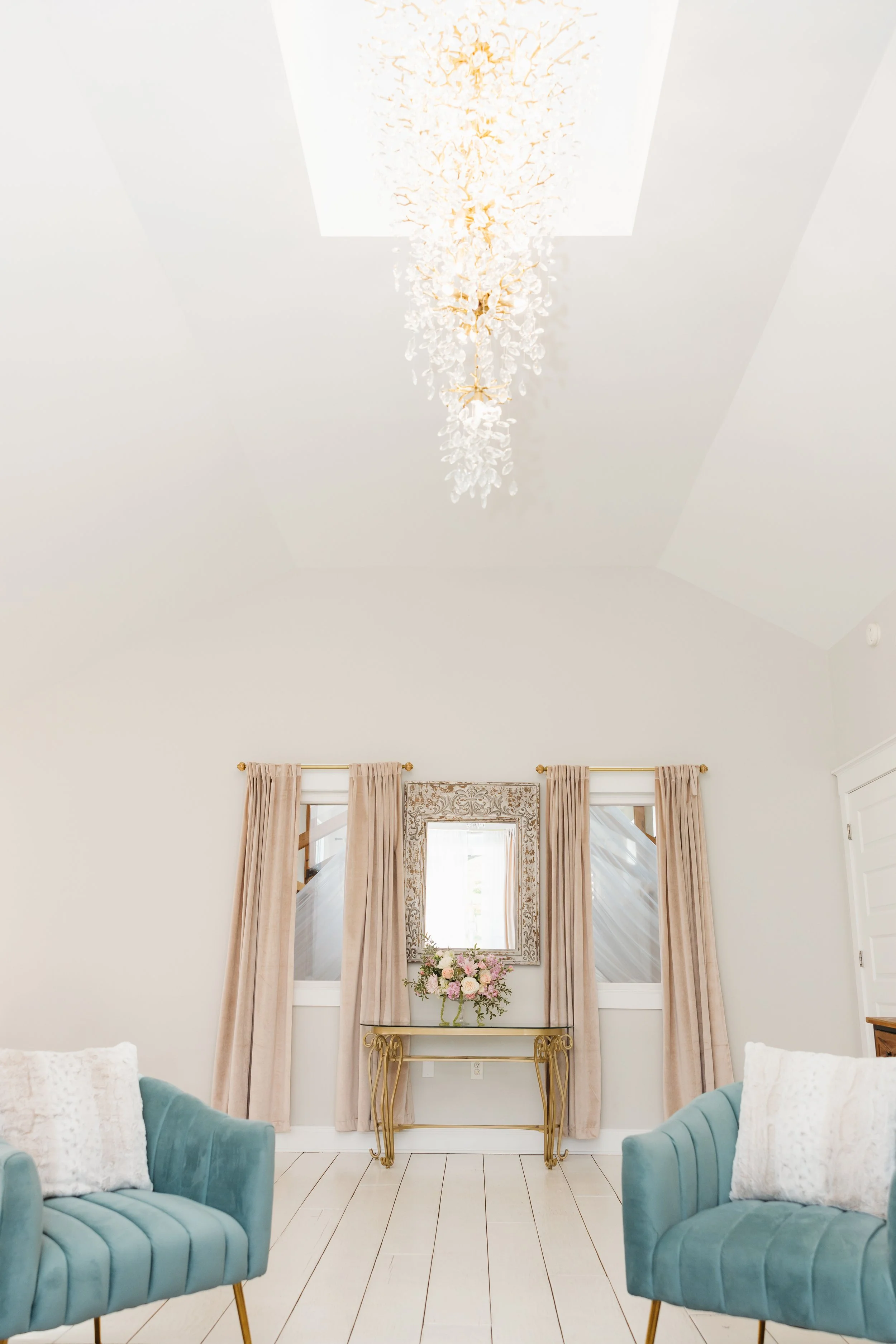 A white bridal suite with a decorative chandelier hanging from the ceiling, beige curtains framing a window with a floral bouquet on a gold table below, and two light blue armchairs with white pillows in the foreground.