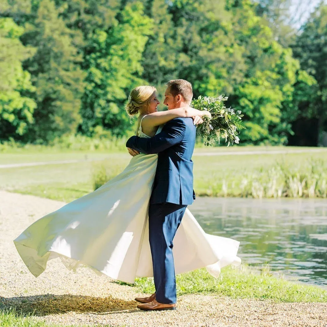 A groom lifting a bride in wedding attire outside near a lake, with green trees in the background.