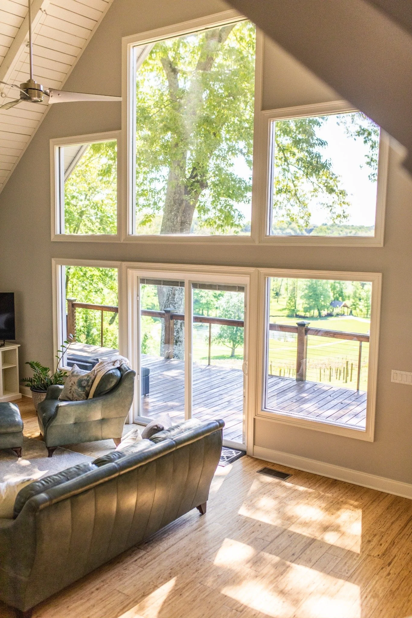 Sunlit living room at the Manor onsite accommodation at Ramble Creek, featuring vaulted ceilings, large windows, leather seating, and doors opening to a wooded deck view