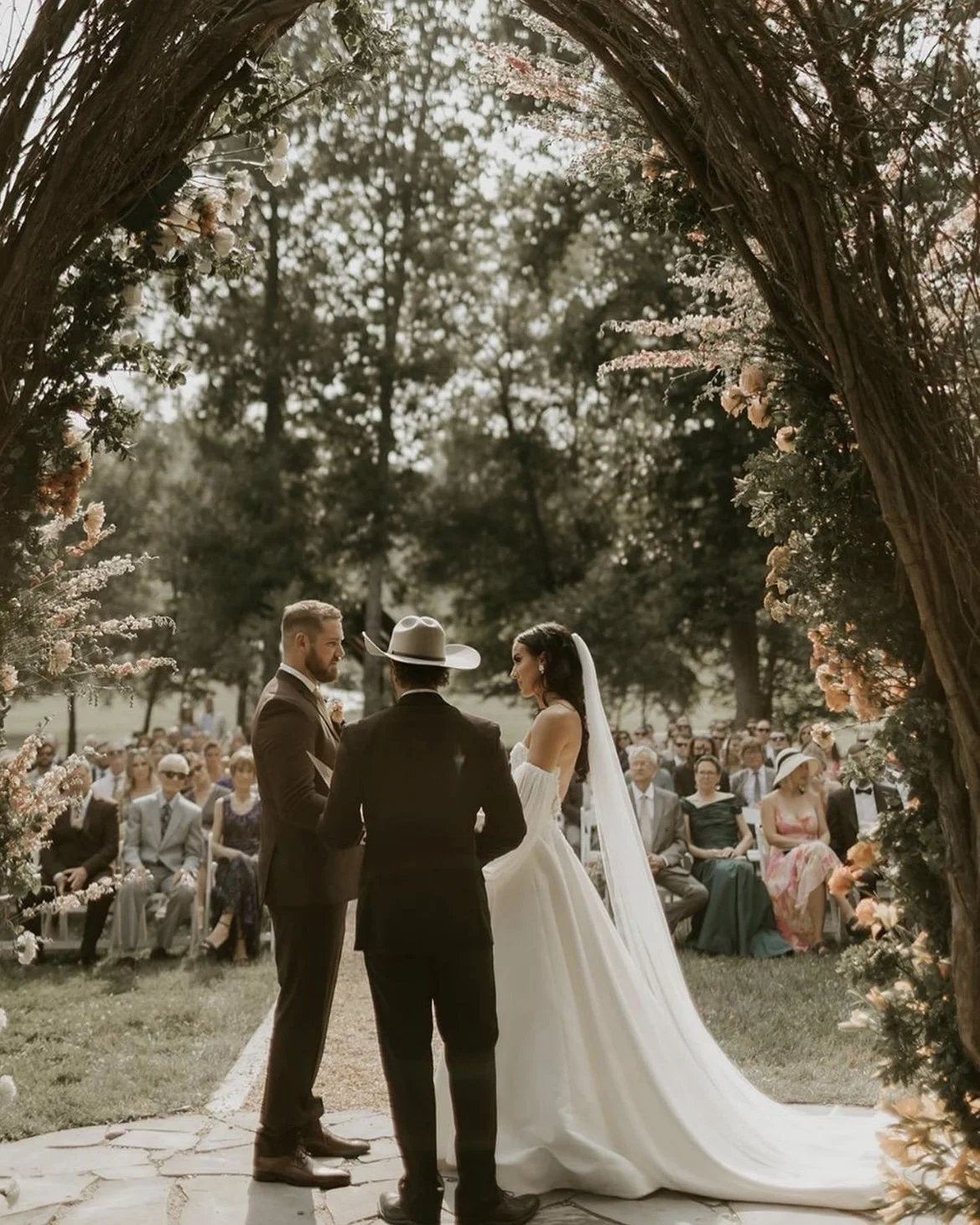 A wedding ceremony taking place outdoors under a floral arch, with the bride and groom facing each other and a officiant in between, surrounded by seated guests.