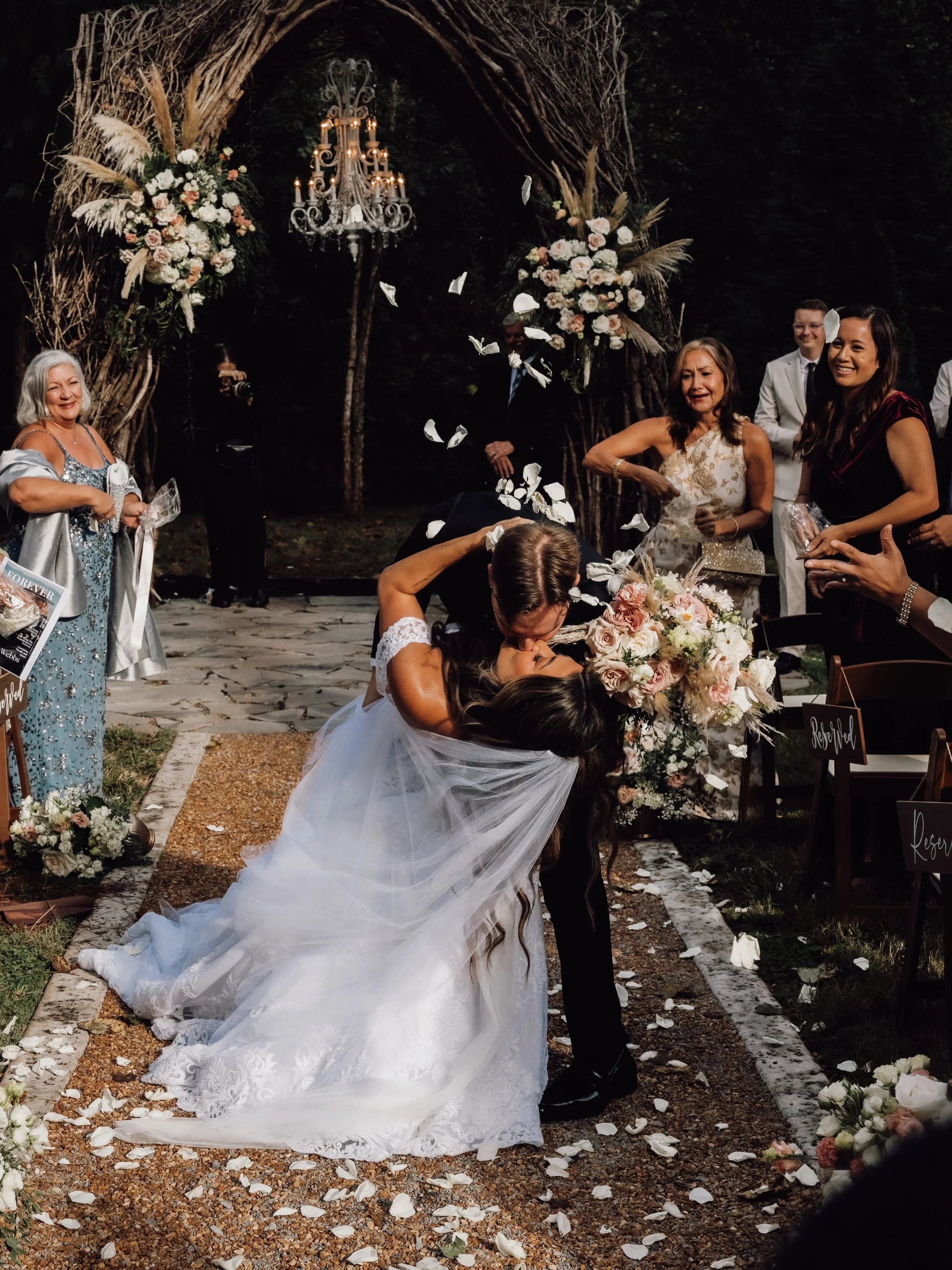 West Webb dips Kara Lewis for a kiss as guests toss petals, with her blush and ivory bouquet and Ramble Creek’s flower-adorned Vine Arbor behind them