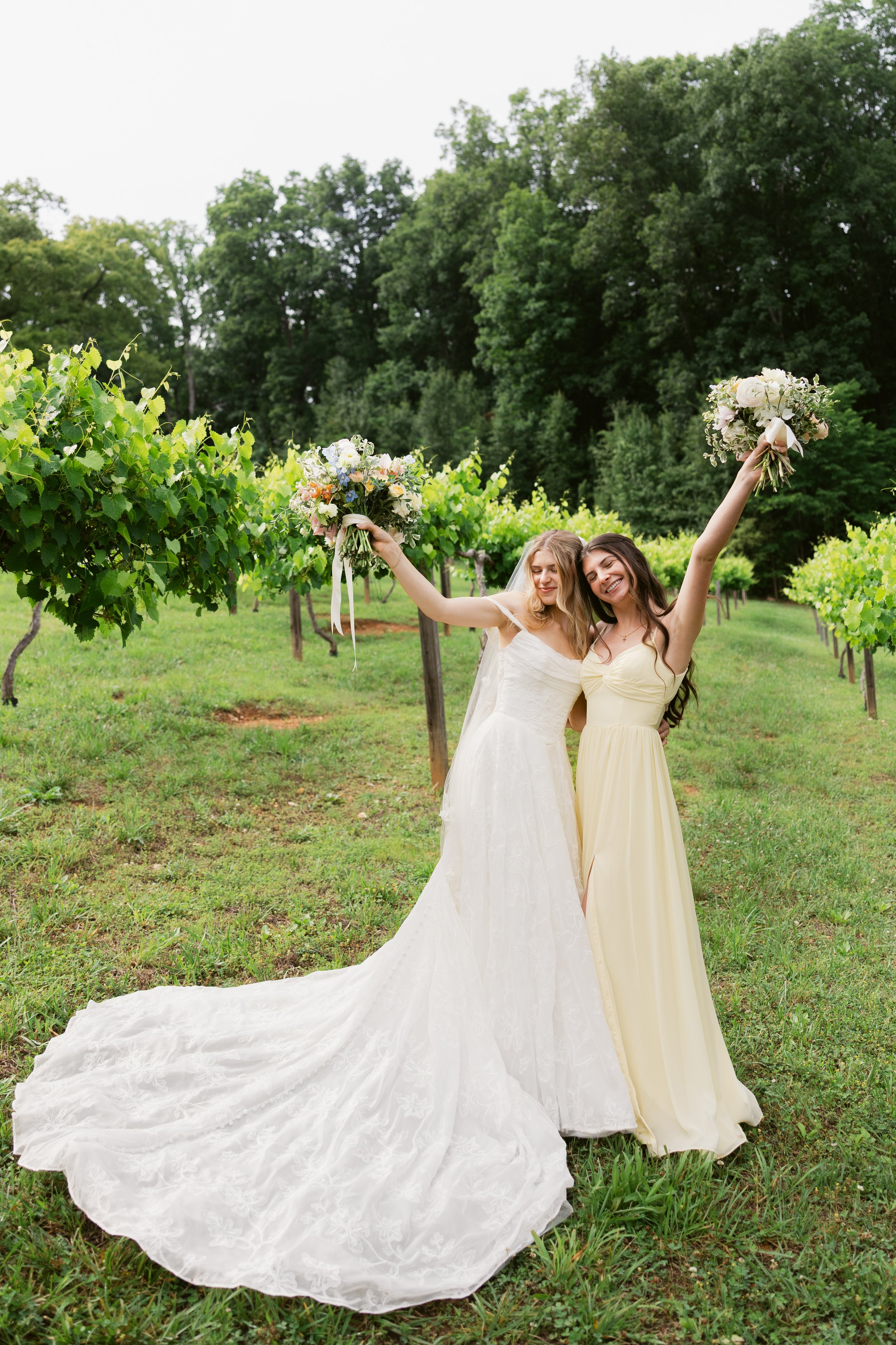Bride and bridesmaid raising bouquets in Ramble Creek’s vineyard, surrounded by lush summer greenery at the Tennessee wedding venue.