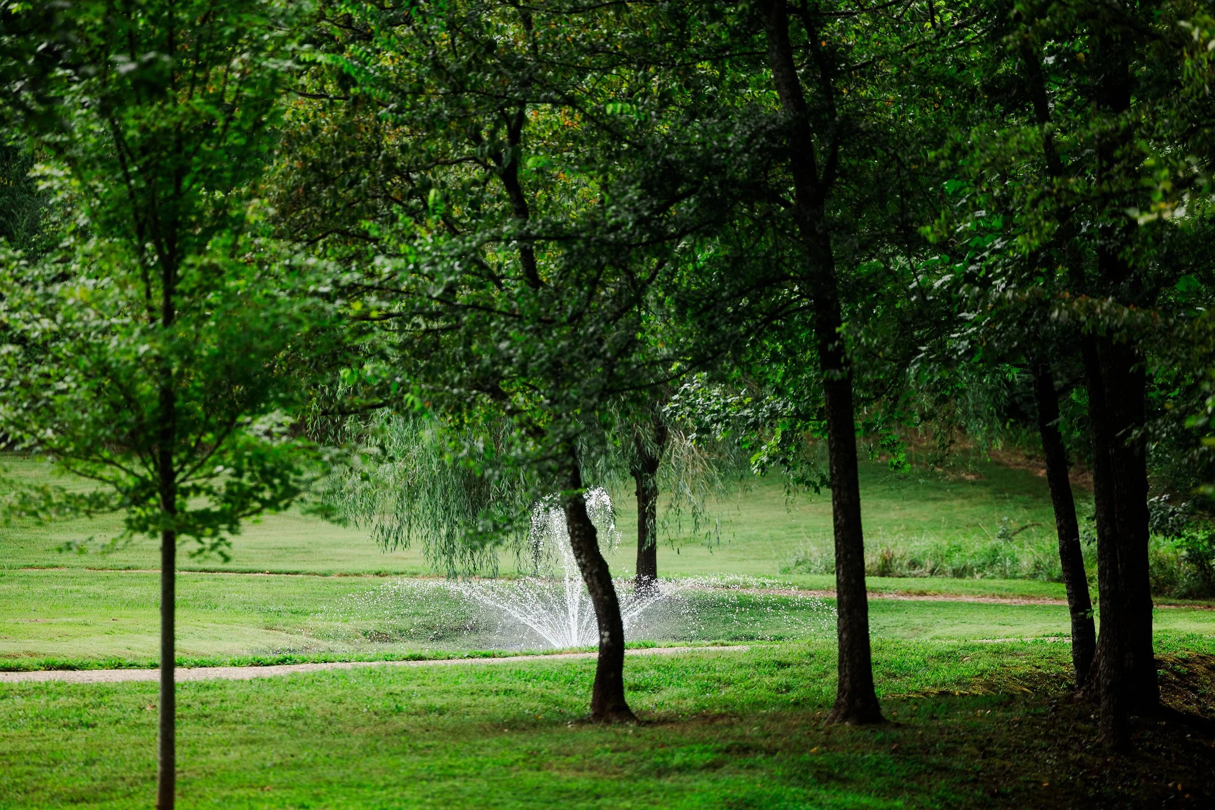 Pond fountain at Ramble Creek surrounded by lush green trees and landscaped grounds during summer at the Tennessee wedding venue.