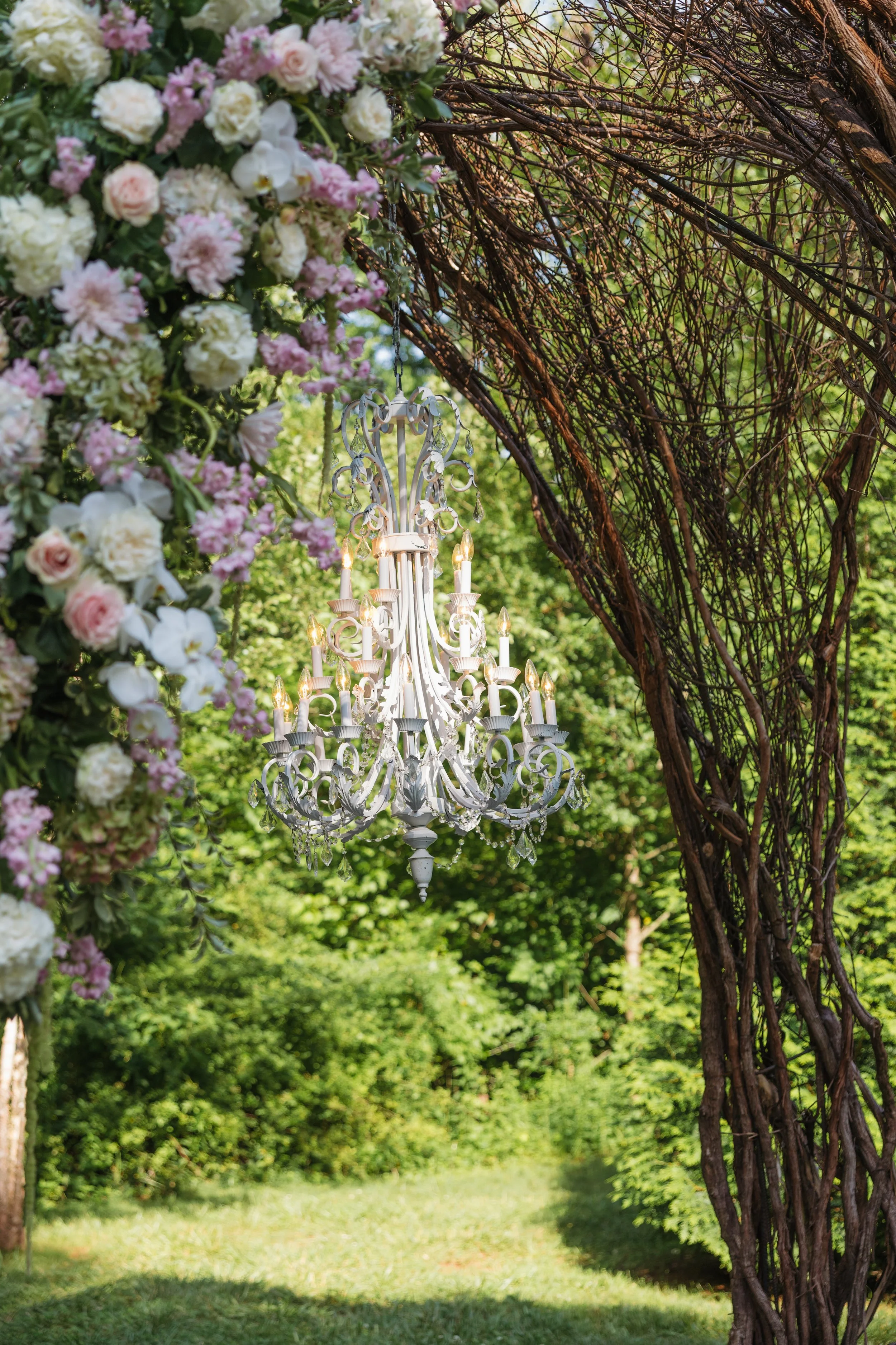 Outdoor ceremony arch at Ramble Creek featuring a hanging chandelier, lush greenery, and romantic blush and ivory floral arrangements