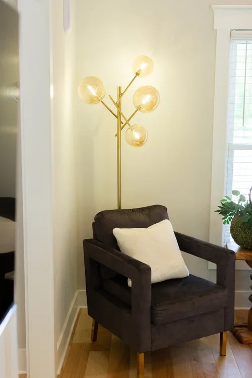 Accent seating nook inside the Villa at Ramble Creek, featuring a dark upholstered chair, white pillow, gold floor lamp, side table, and window.
