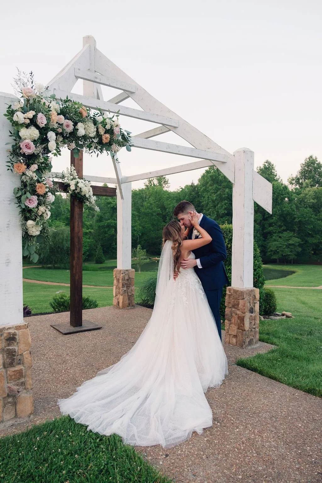 Bride and groom kissing beneath a white outdoor wedding arbor with a floral-covered cross, stone columns, and green lawns surrounding the ceremony space