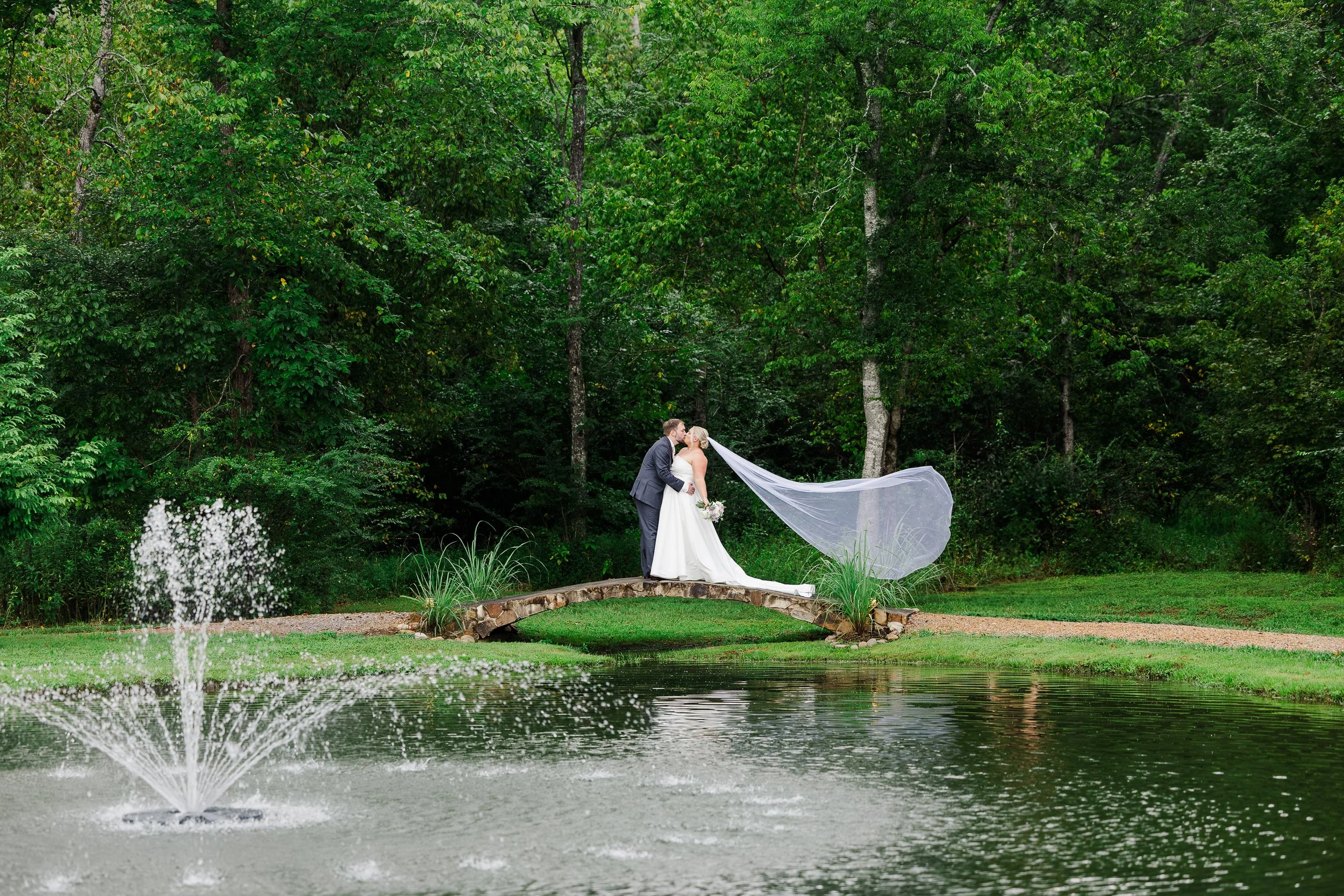 Bride and groom kissing on Ramble Creek’s stone bridge by the pond fountain, surrounded by lush Tennessee greenery during a summer wedding.