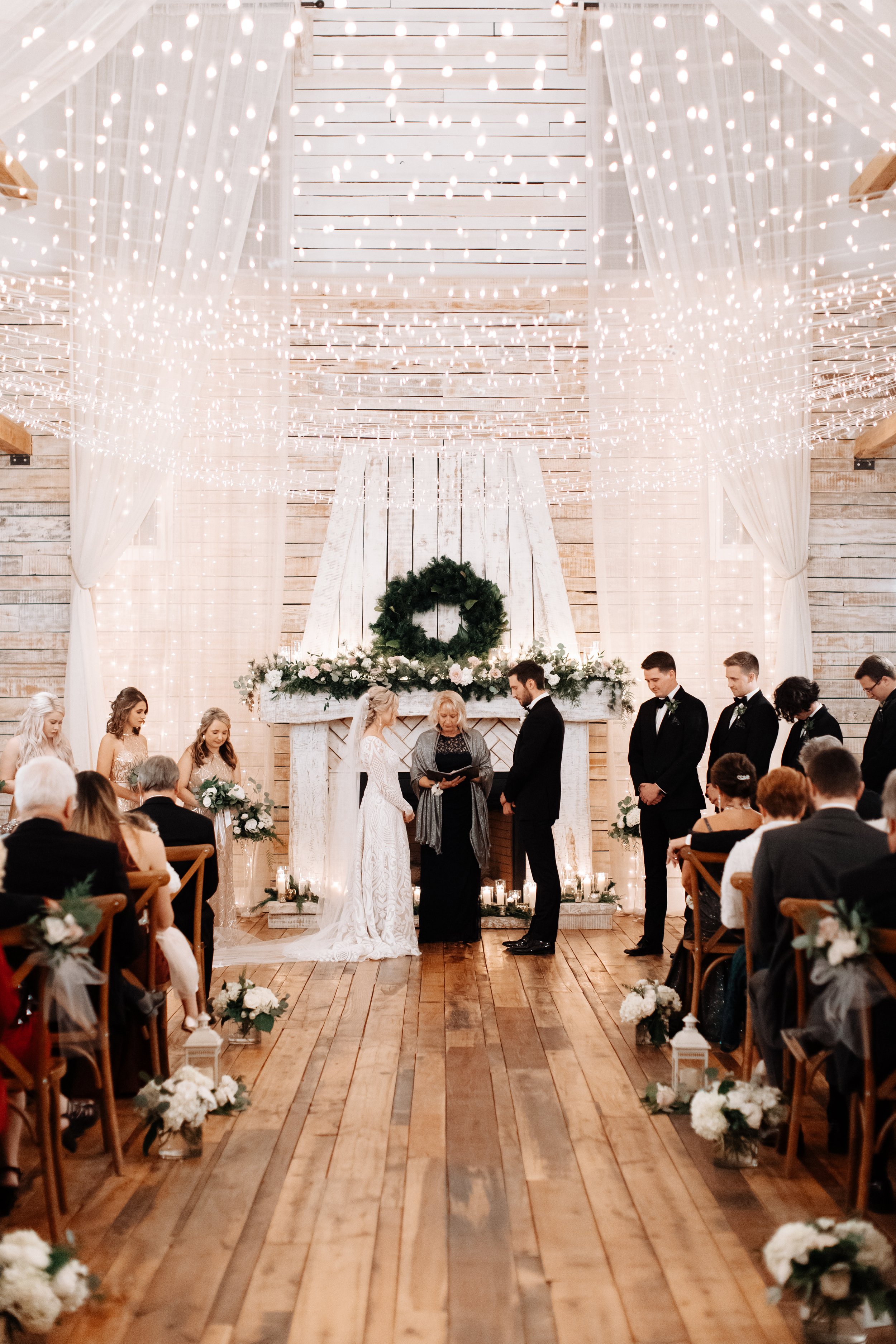 An indoor wedding ceremony in a white, airy, light-filled wedding venue with string lights, draped curtains, and floral arrangements. The bride and groom are standing facing each other, with the officiant in between, surrounded by friends and family.