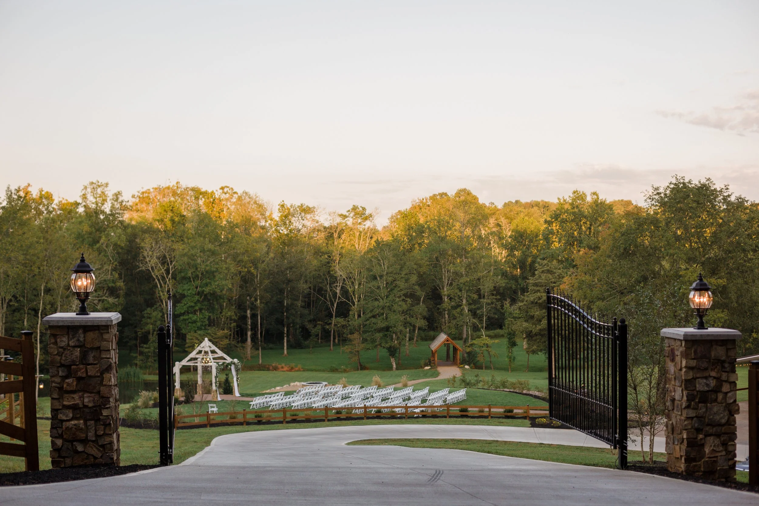 View of a large open lawn area with white chairs arranged in rows, a white gazebo, and trees in the background, seen through black iron gates with stone pillars and lamps on either side at Ramble Creek wedding venue in Athens, TN.