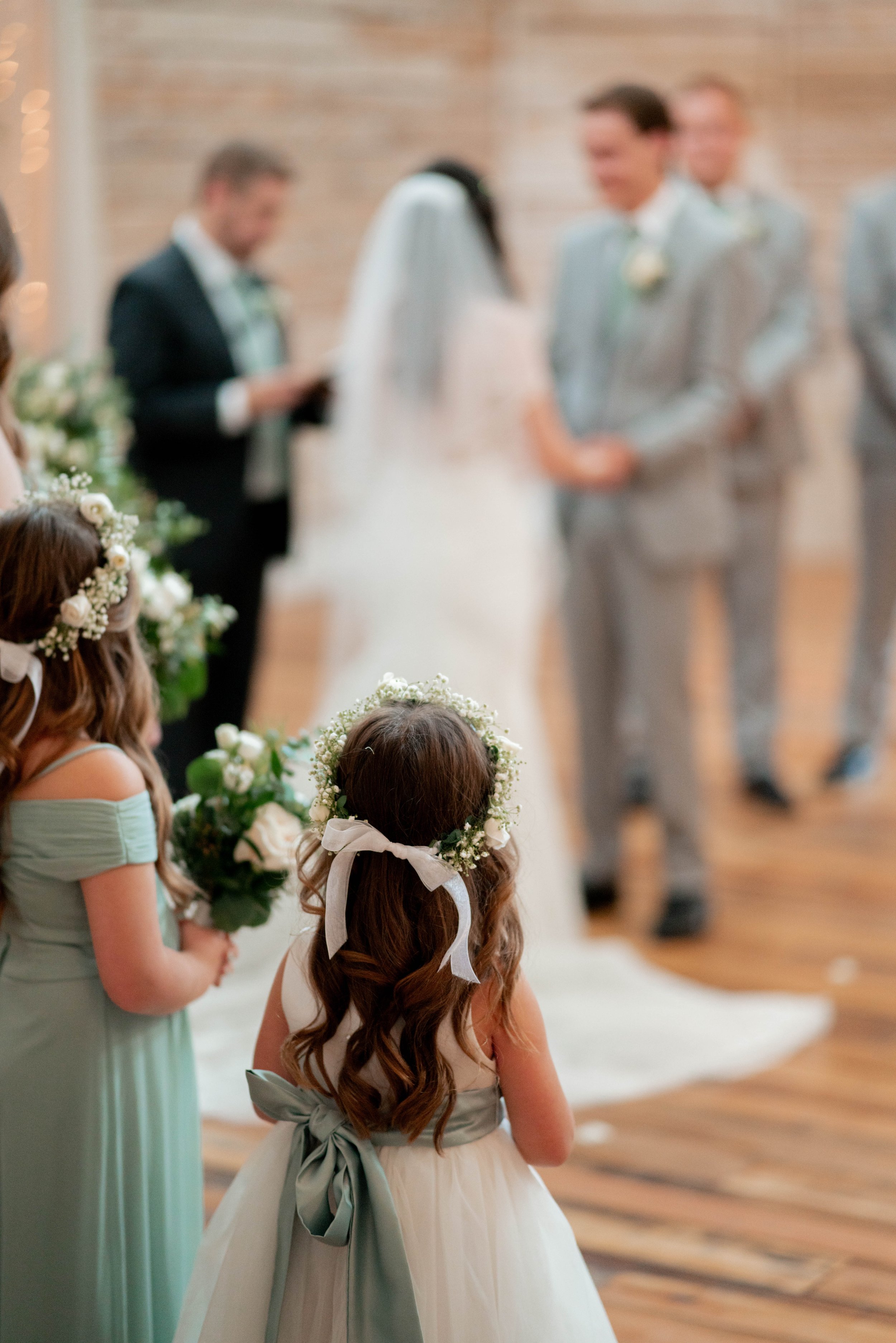 Children and adults in formal attire participating in a wedding ceremony indoors at Ramble Creek. Little girls are holding bouquets and wearing floral crowns. The bride and groom are facing each other in the background.